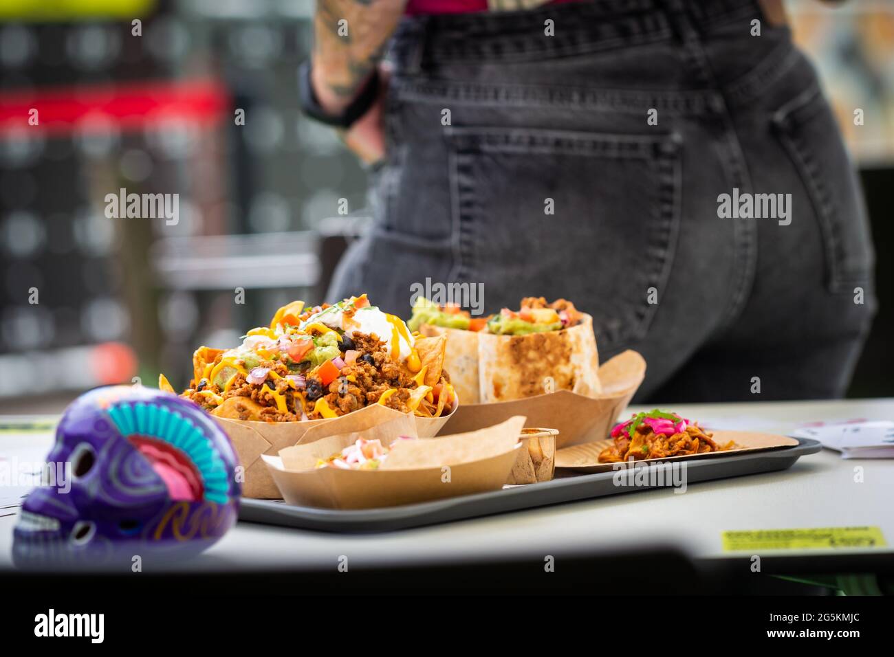 Un uomo si leva in piedi cibo messicano tradizionale in un caffè fast food con un cranio al tavolo in background. Burritos, salsa nachos e salse Foto Stock
