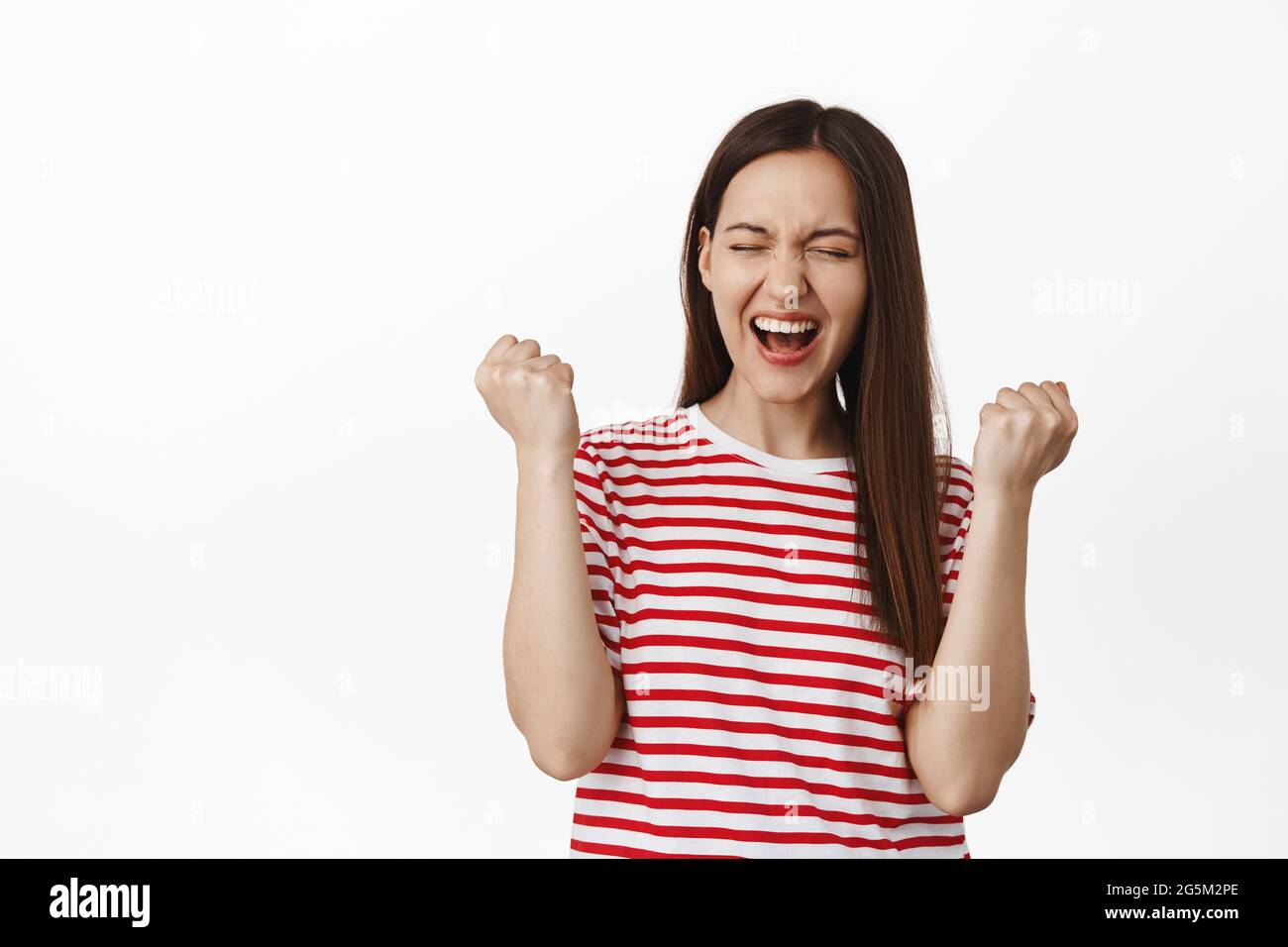 Immagine di una giovane donna entusiasta pompa pugno, ragazza gridando sì e trionfando, raggiungere il traguardo, celebrare la vittoria, in piedi in t-shirt contro bianco Foto Stock