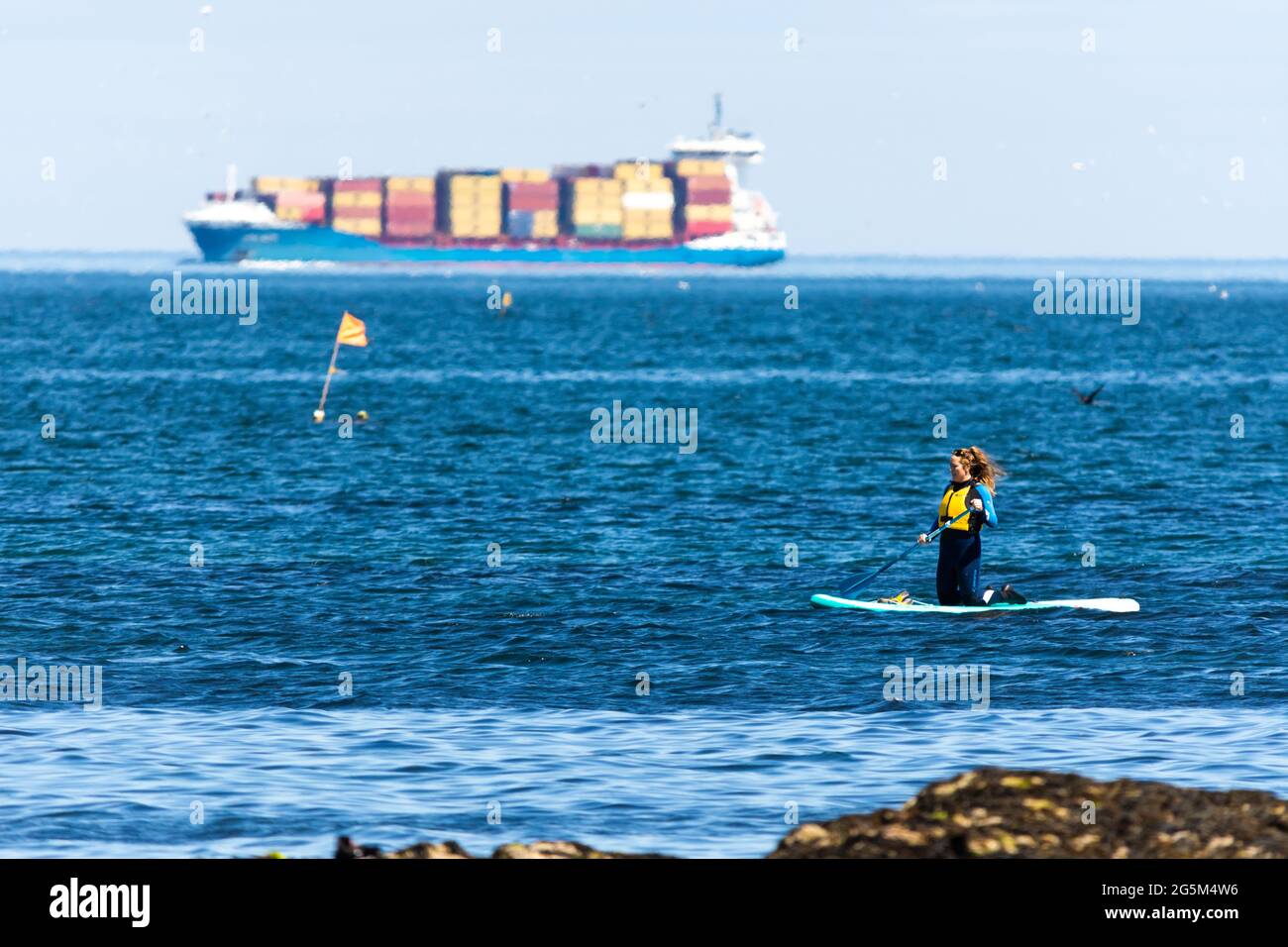Un'ignota pagaia è salita a bordo delle pale nel firth of Forth, in Scozia, mentre un'enorme nave portacontainer passa sullo sfondo Foto Stock