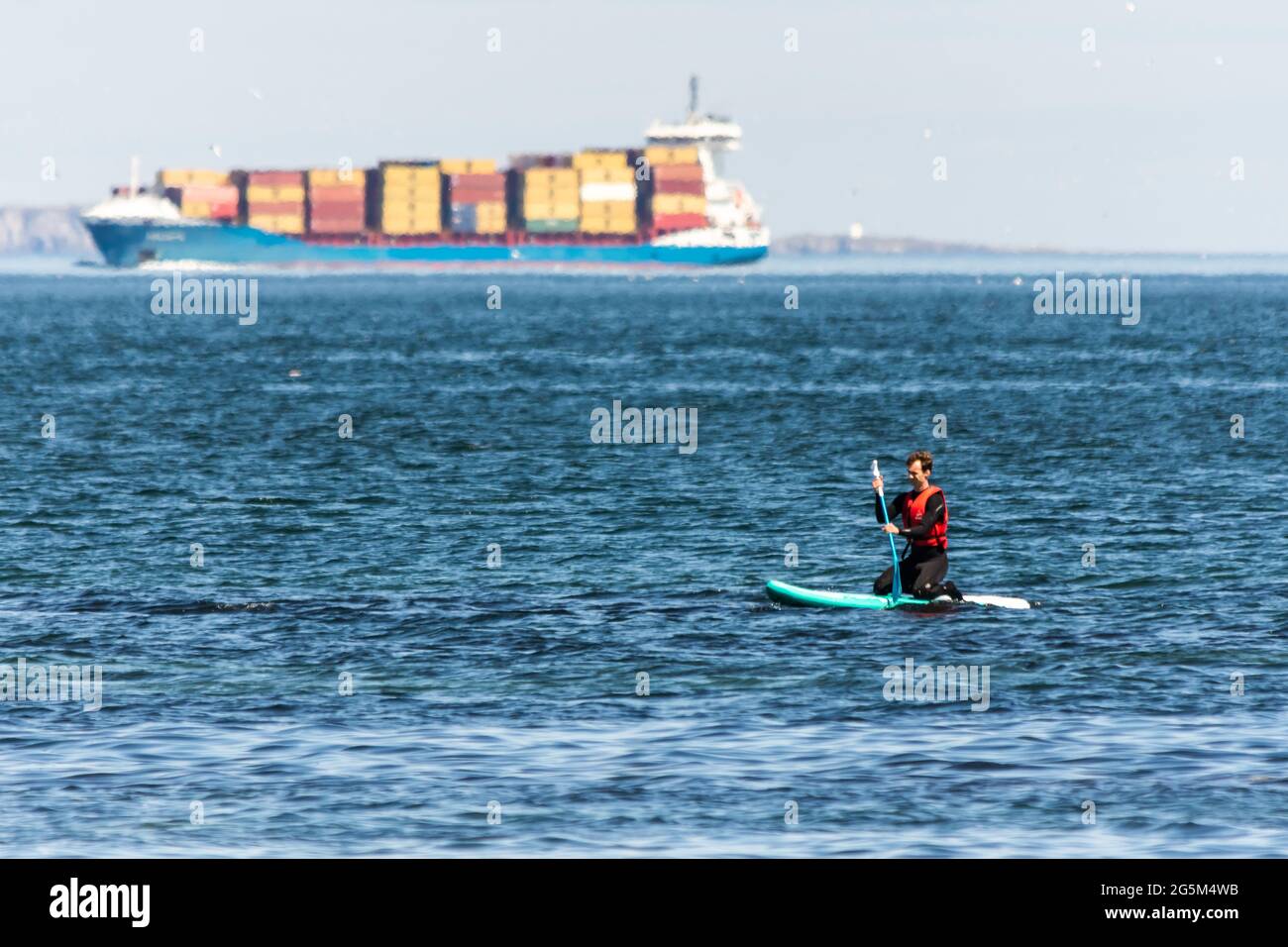 Un'ignota pagaia è salita a bordo delle pale nel firth of Forth, in Scozia, mentre un'enorme nave portacontainer passa sullo sfondo Foto Stock