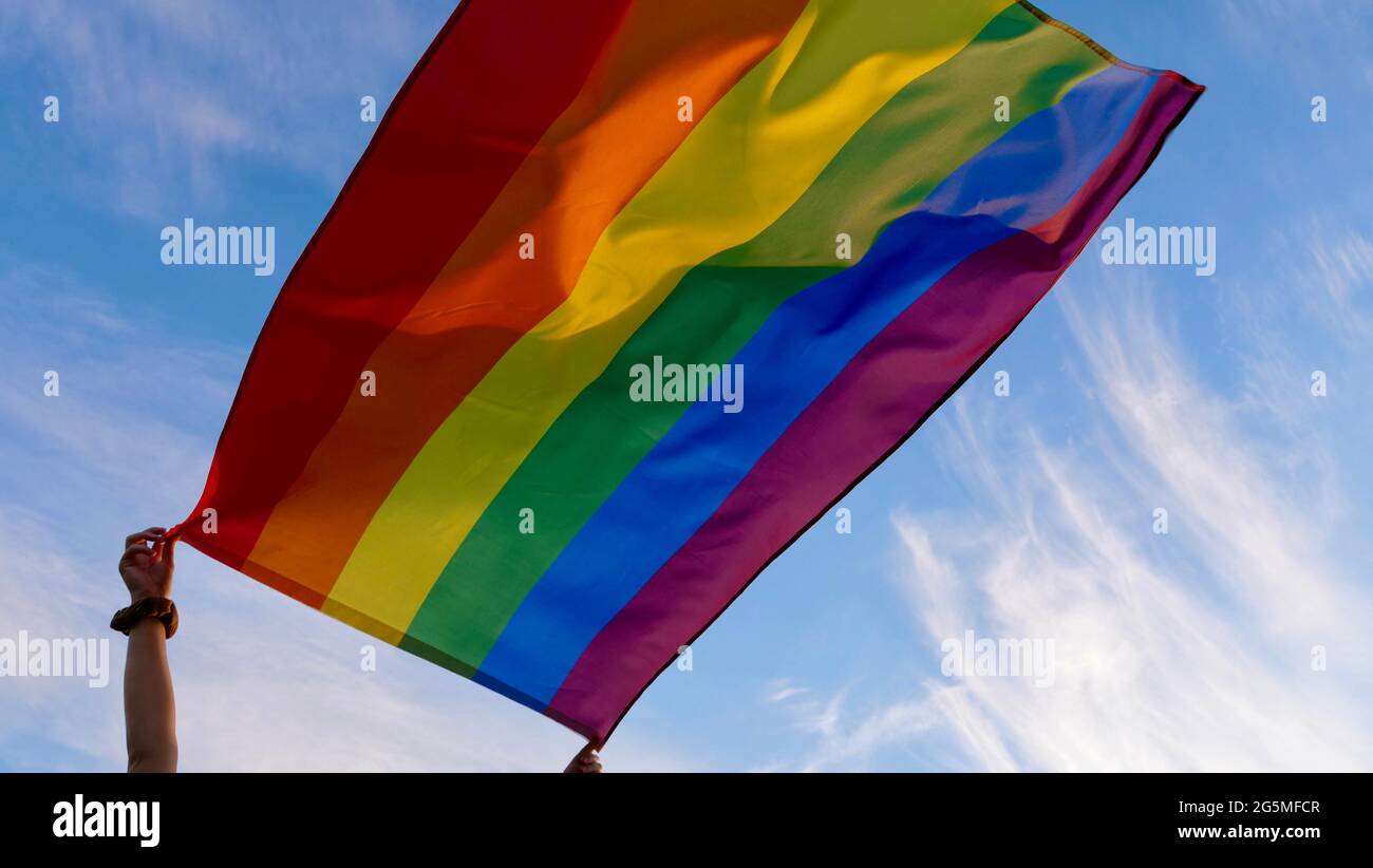 Mano femminile che tiene una bandiera di identità di genere LGBT su uno sfondo di cielo blu nuvoloso in un sole ventoso giorno estivo durante la celebrazione di una parata gay, bisessualità o uscire giorno Foto Stock