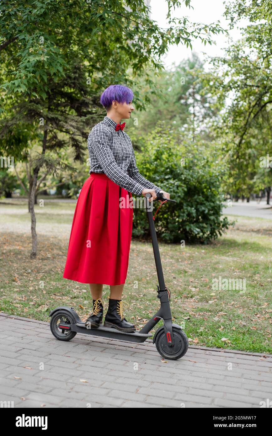 Una giovane donna con i capelli viola guida uno scooter elettrico in un  parco. Ragazza elegante in una camicia a plaid, una lunga gonna rossa e una  cravatta di prua sta cavalcando