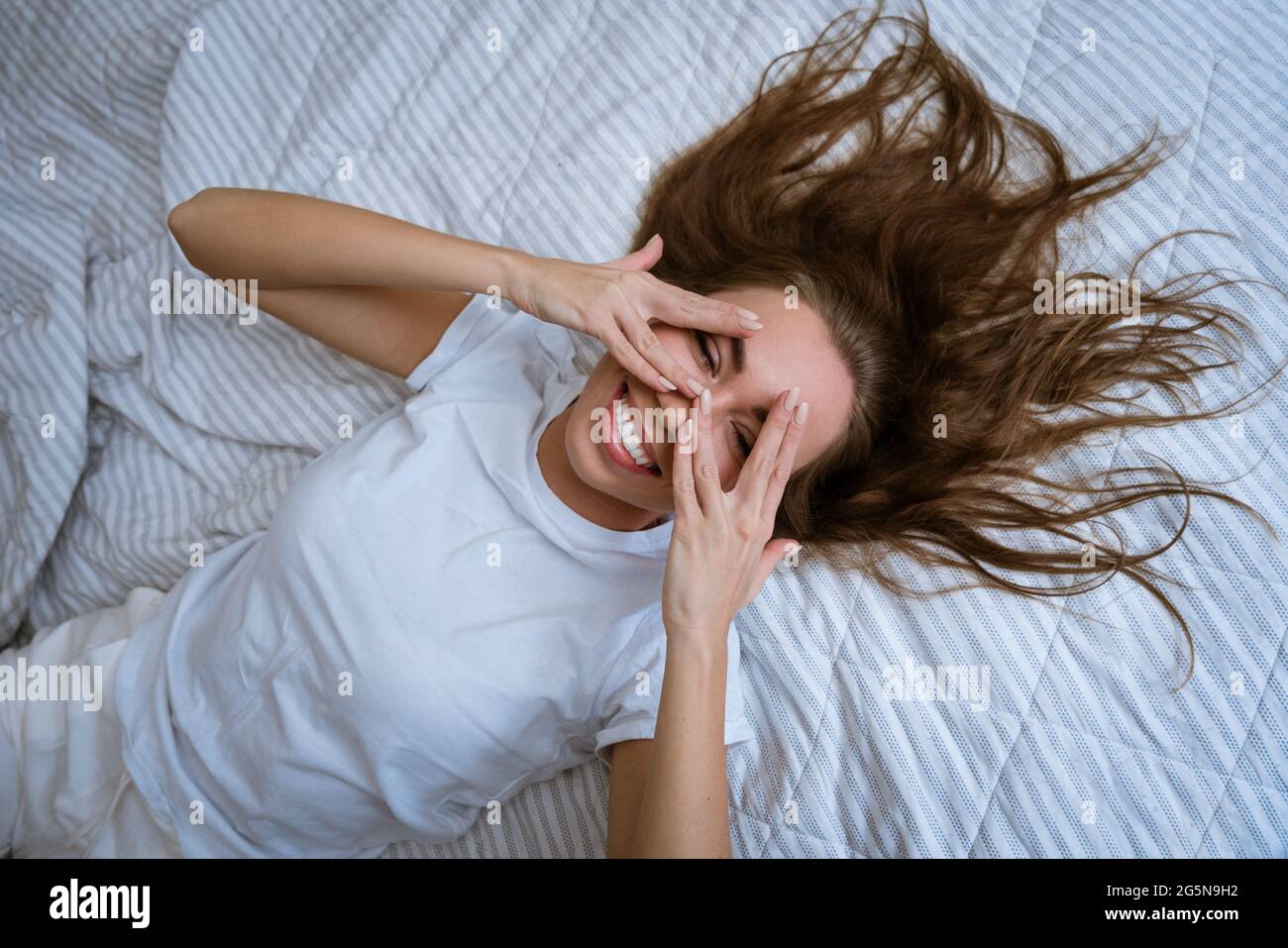 Bella giovane donna sdraiata a letto sorridente. Una ragazza con gli occhi  aperti copre il viso con le mani e le dita al mattino. Concetto di ragazza  felice. Vista dall'alto. La gioia