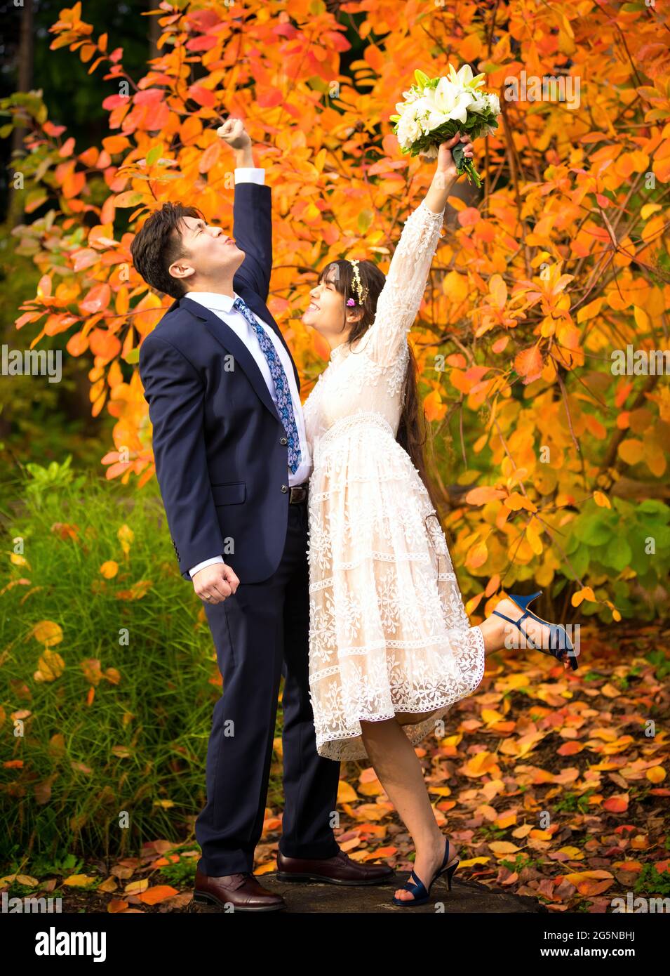 Sposo e sposo biraciale appena sposato sollevando le braccia che si acclamano dopo il matrimonio mentre si levano in piedi all'aperto in autunno, foglie di arancio luminoso in background Foto Stock