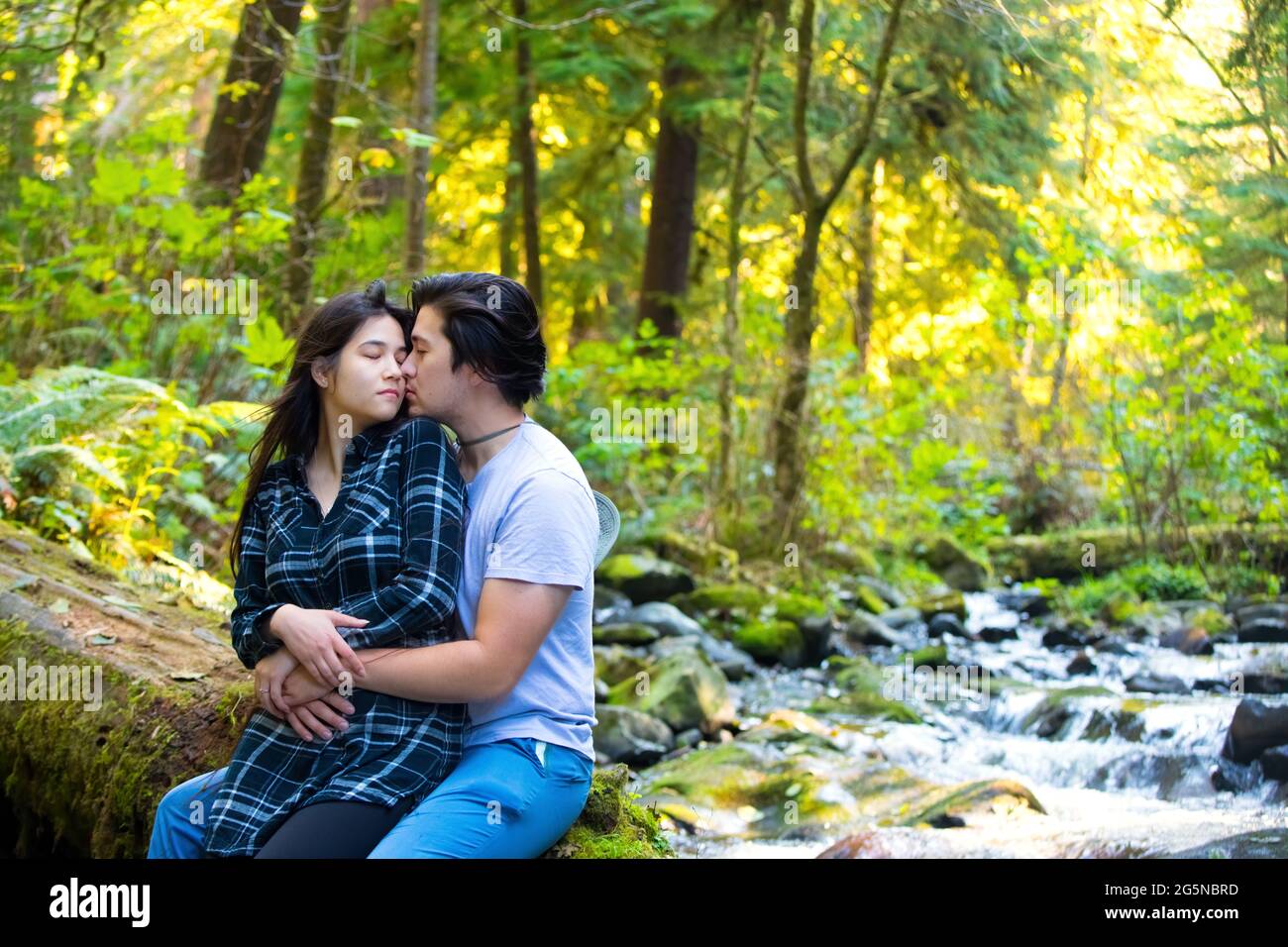 Giovane coppia biraciale seduta lungo il ruscello roccioso in boschi verdi che abbracciano Foto Stock