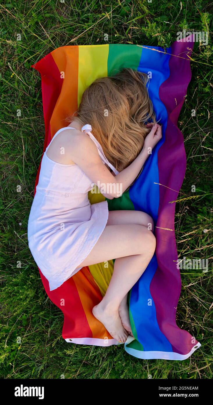 Donna lesbica sdraiata sulla bandiera LGBT sull'erba in abito bianco. Sostegno all'orientamento non tradizionale nel mese della dignità, giorno della bisessualità. La persona perde e nasconde il suo volto dal bullismo. Foto Stock