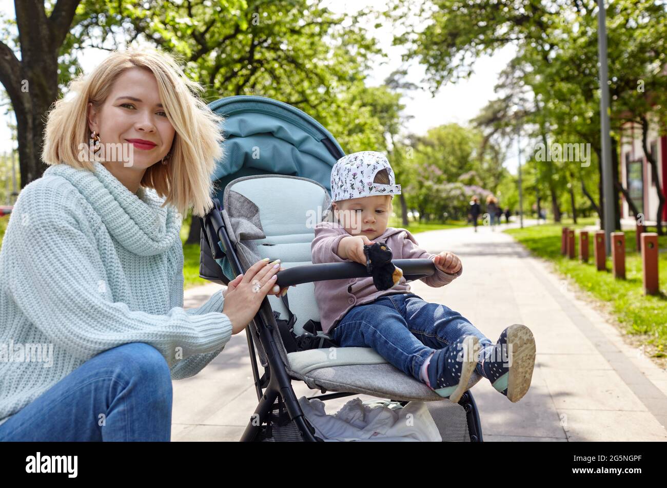 Mamma e figlia sulla natura a piedi presso il parco primaverile. Adorabile bambina in abiti comuni seduta in carrozzina blu. Bambino in buggy Foto Stock