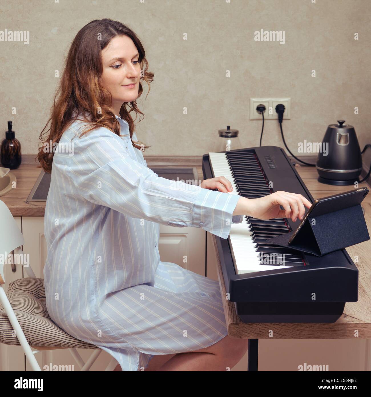 Una donna in una notte guarda la musica su un tablet digitale e suona il pianoforte di casa. Imparare la musica online nel periodo della pandemia in isolamento Foto Stock