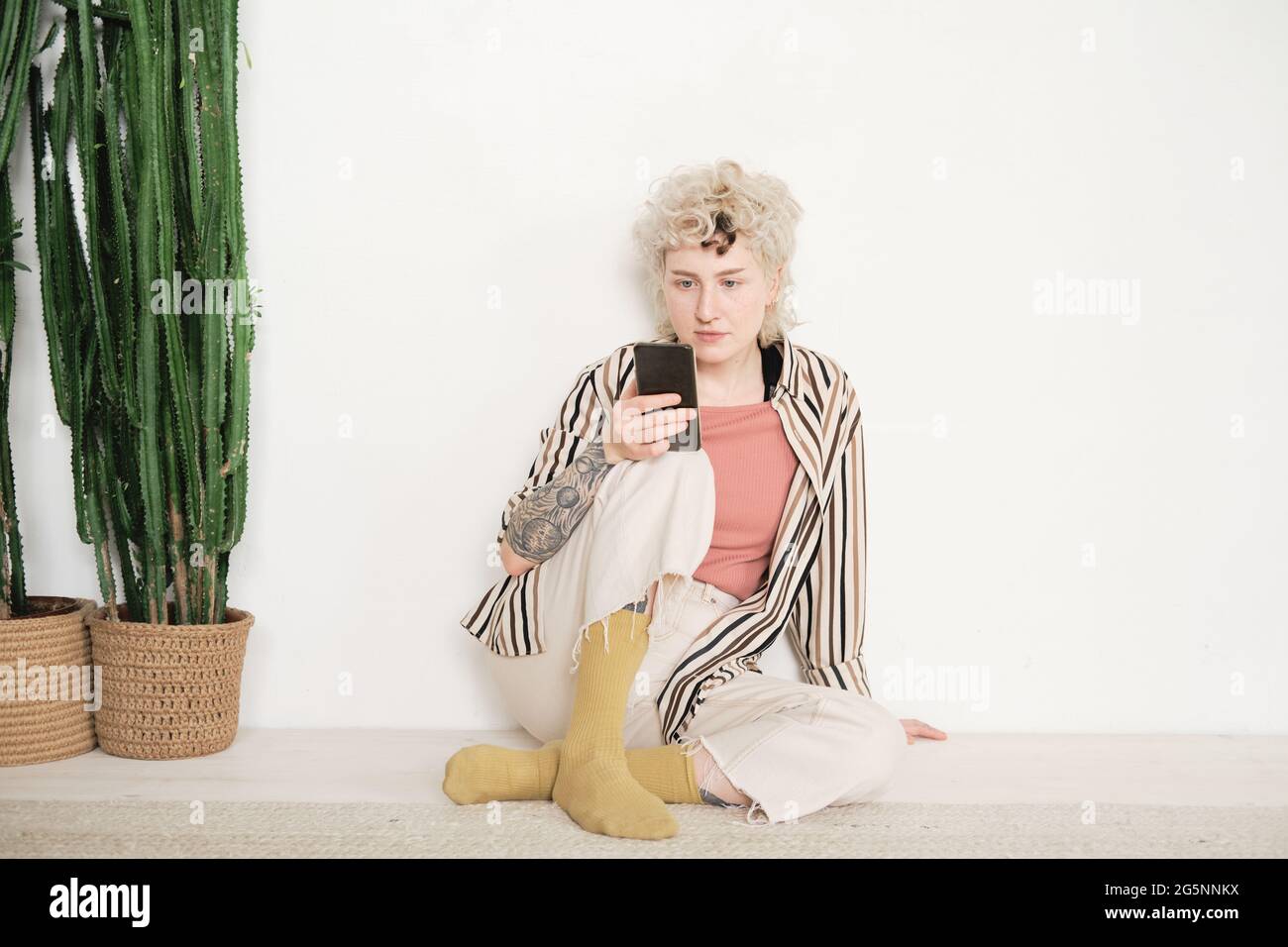 Giovane donna elegante seduta sul pavimento e leggendo online sul telefono cellulare Foto Stock