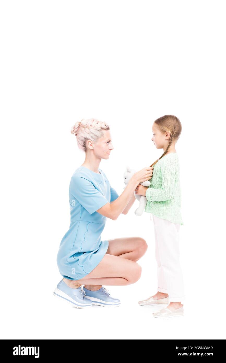 Scatto isolato verticale di lunghezza intera di madre moderna seduta davanti a sua figlia fissando i suoi capelli, sfondo bianco Foto Stock
