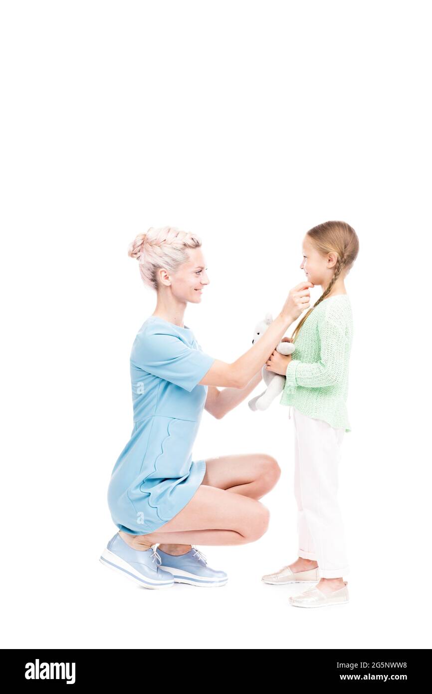 Scatto isolato verticale di lunghezza intera di madre moderna seduta davanti a sua figlia che tocca il mento, sfondo bianco Foto Stock