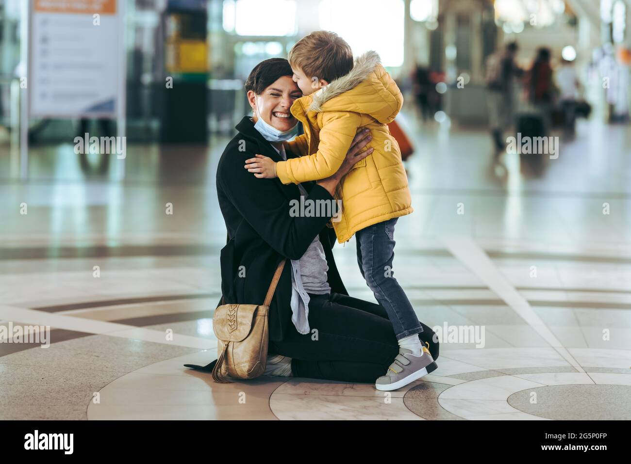 Madre che indossa la maschera di contatto con suo figlio dopo l'arrivo da viaggio di lavoro in aeroporto. Capretto che ama la sua madre che si siede sul pavimento all'aeroporto. Foto Stock