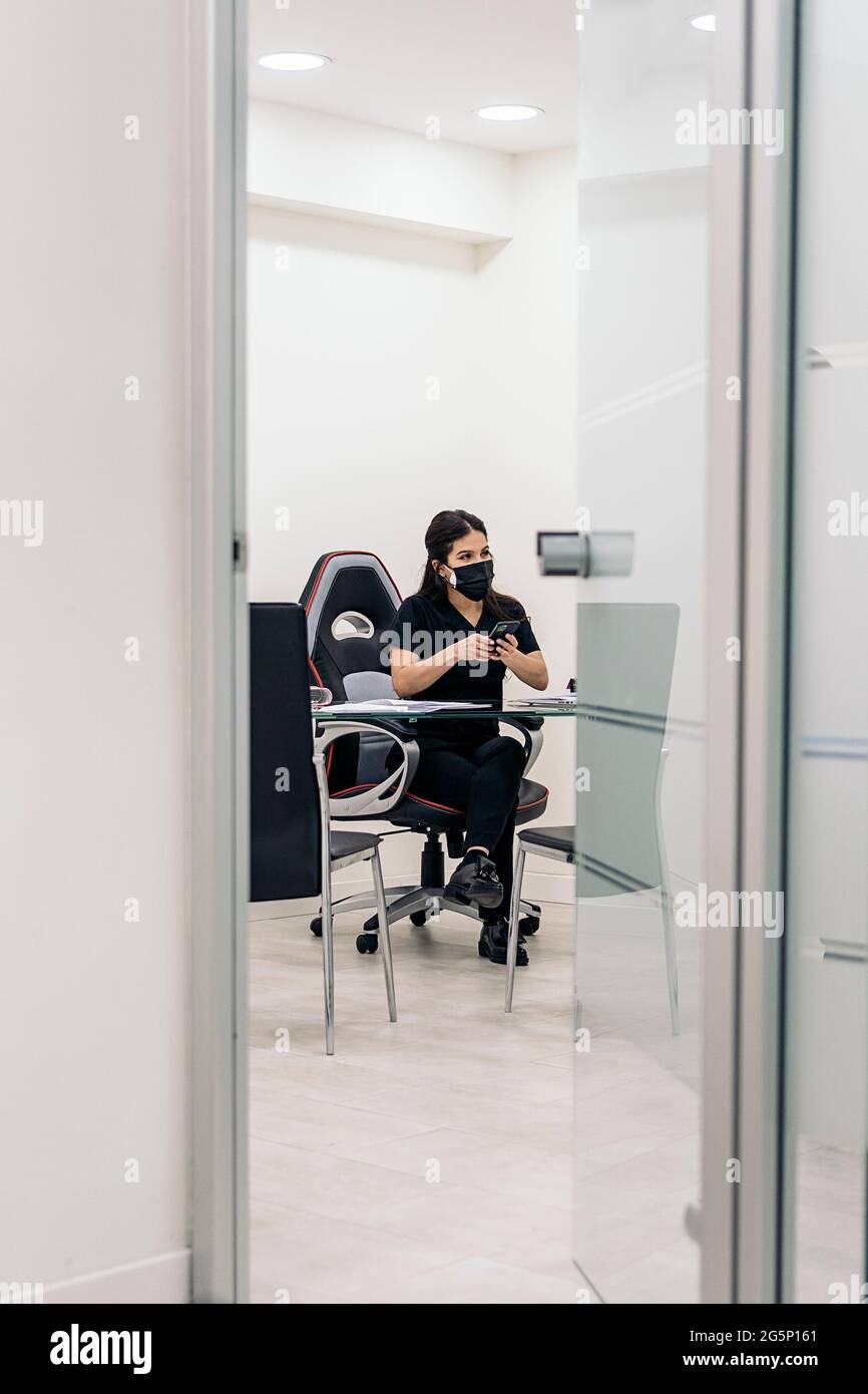 Giovane dentista donna che indossa la maschera per il viso a causa di covid19 che lavora nel suo ufficio e utilizzando il suo telefono. Foto Stock