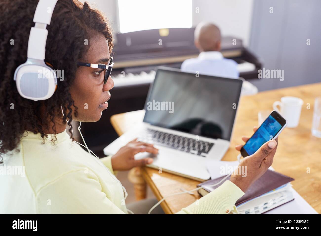 Donna africana con cuffia che ha una chat video in ufficio domestico con bambino al pianoforte in background Foto Stock
