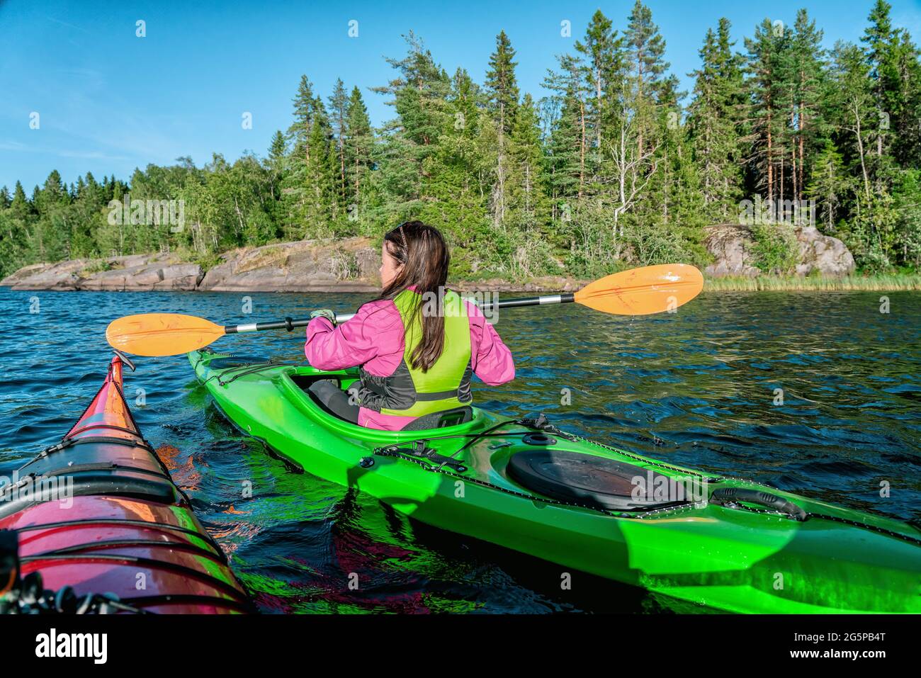 Donne in kayak che tengono le pagaie orizzontalmente mentre passano vicino un'altra persona in kayak. Indossa una giacca magenta e un giubbotto di sicurezza verde. Pino estivo t Foto Stock