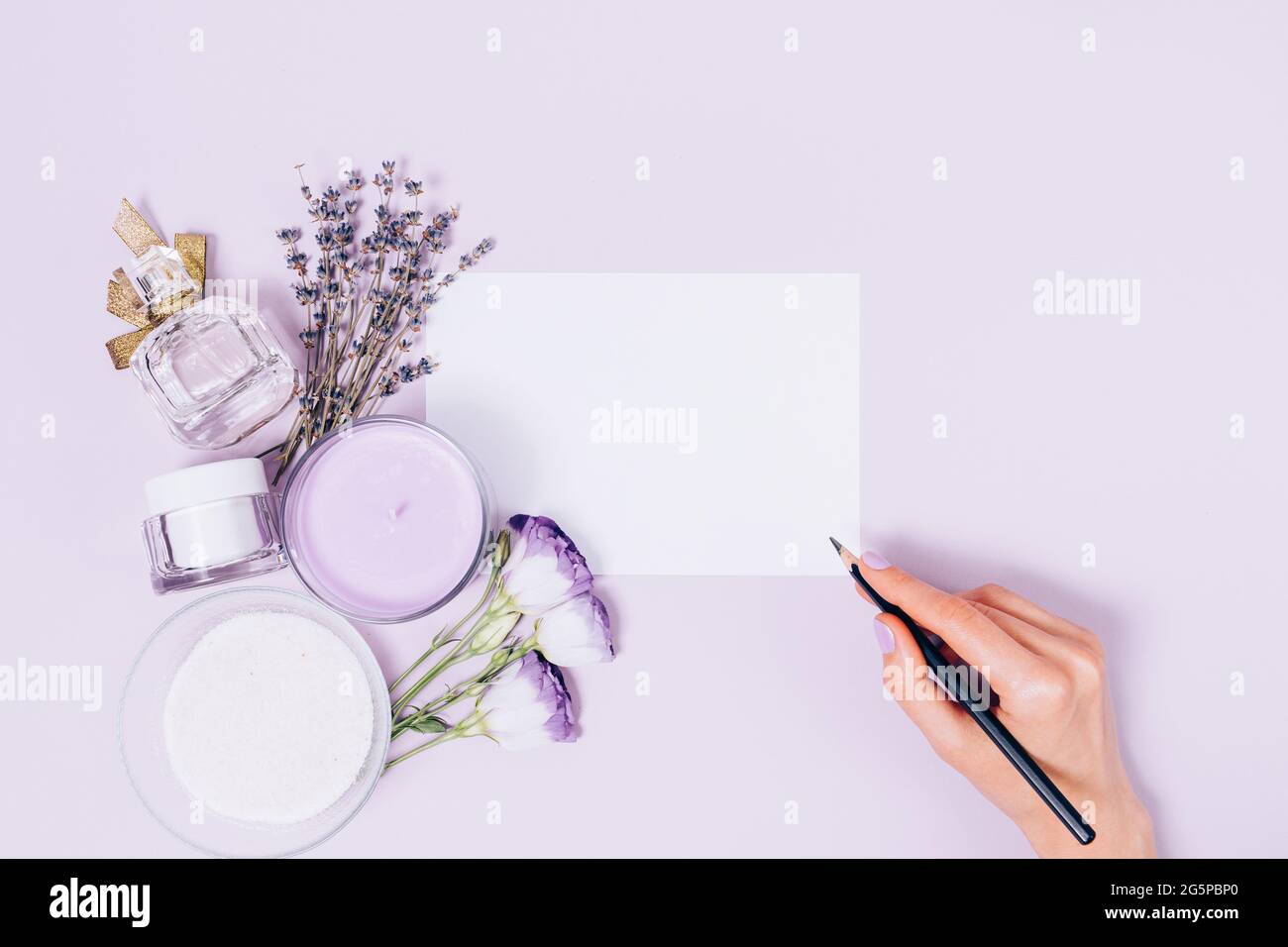 Vista dall'alto della mano femminile che tiene la matita scrive sulla mascherina della scheda vuota vicino ai fiori, ai cosmetici naturali e alla candela profumata sul backgroun chiaro della lavanda Foto Stock