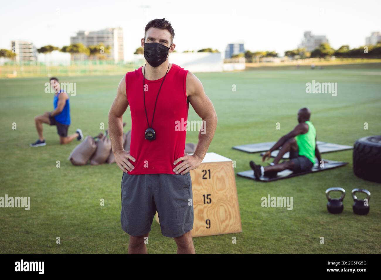 Ritratto di allenatore di fitness maschile caucasico con maschera facciale, con colleghi che si esercitano all'aperto Foto Stock