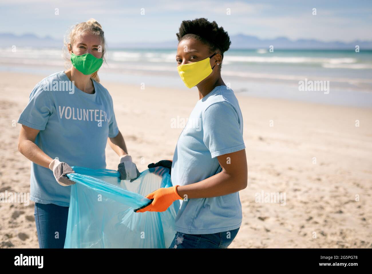 Due donne diverse che indossano t-shirt volontario e maschere facciali che raccolgono rifiuti dalla spiaggia Foto Stock