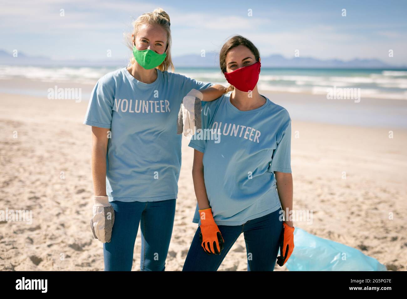 Due donne diverse che indossano t-shirt volontario e maschere facciali che raccolgono rifiuti dalla spiaggia Foto Stock