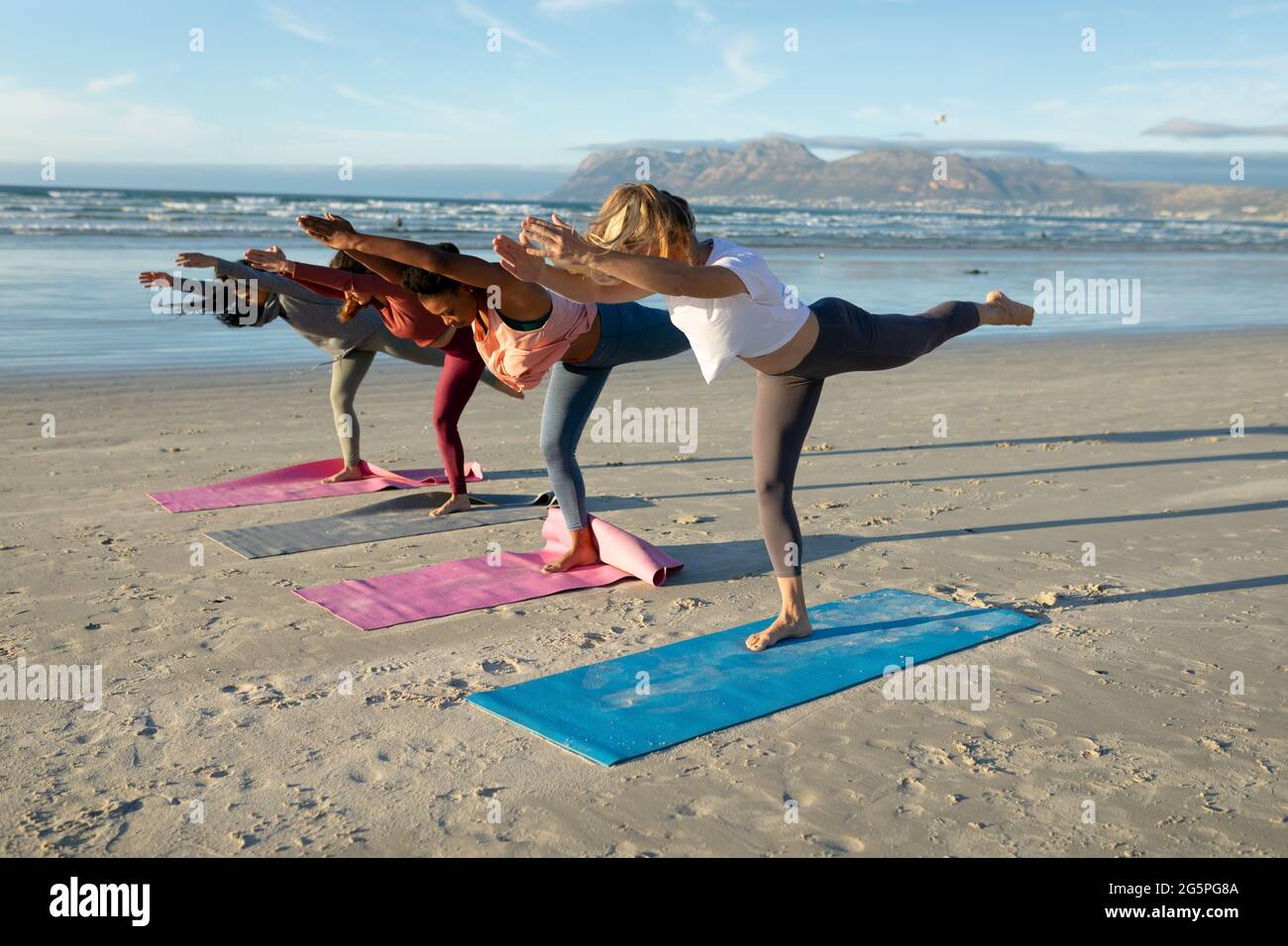 Gruppo di diverse amiche che praticano lo yoga, alzando una gamba che si allunga in spiaggia Foto Stock