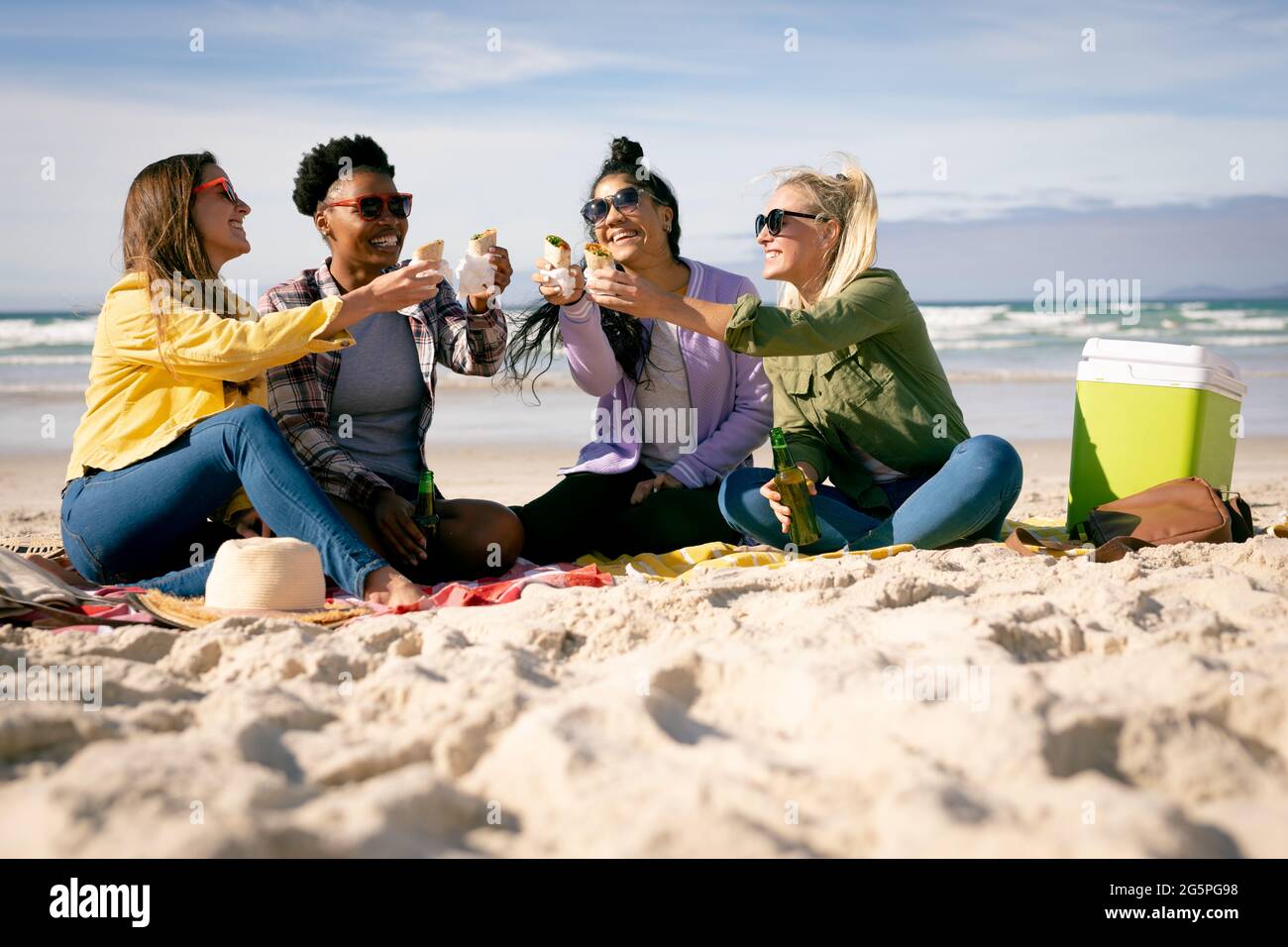 Felice gruppo di diverse amiche che si divertono, sedersi sulla spiaggia tenendo il cibo ridendo Foto Stock