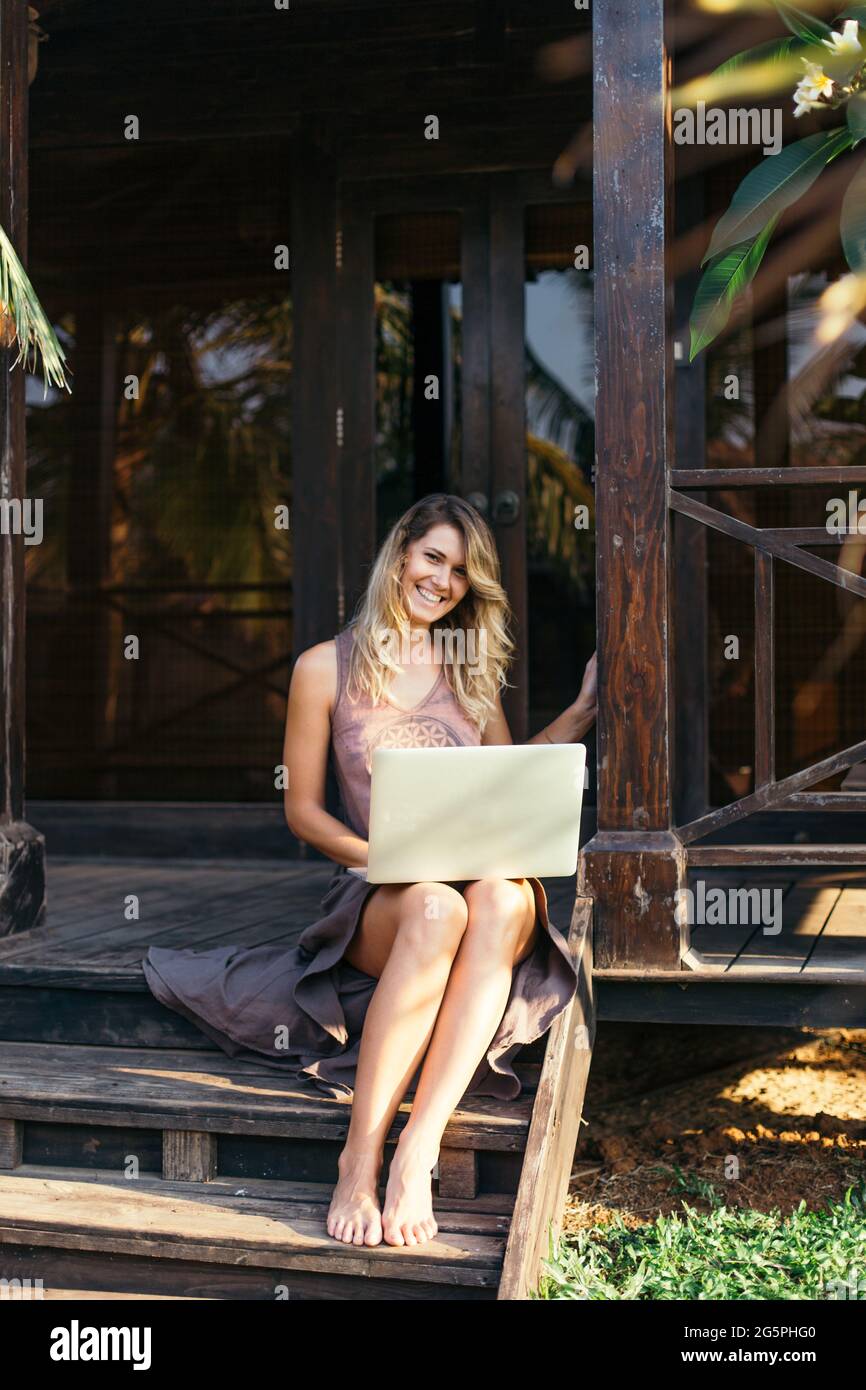 La giovane donna moderna ride felicemente guardando l'atcamera. È seduta su portico di legno di capanna tropicale con il laptop in grembo. Ricreazione e turismo. REM Foto Stock