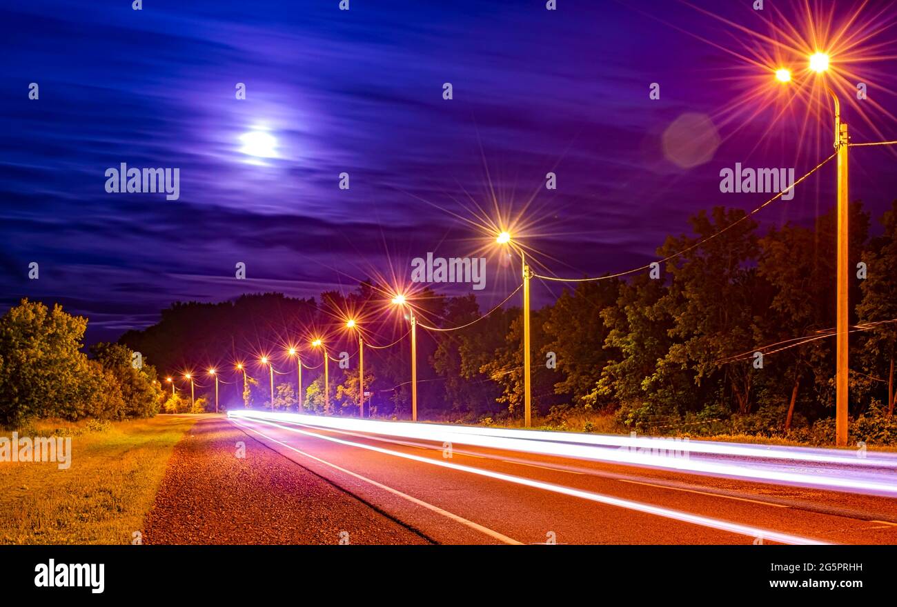 Una lunga linea dai fari di un'auto che passa su una strada asfaltata notturna Foto Stock