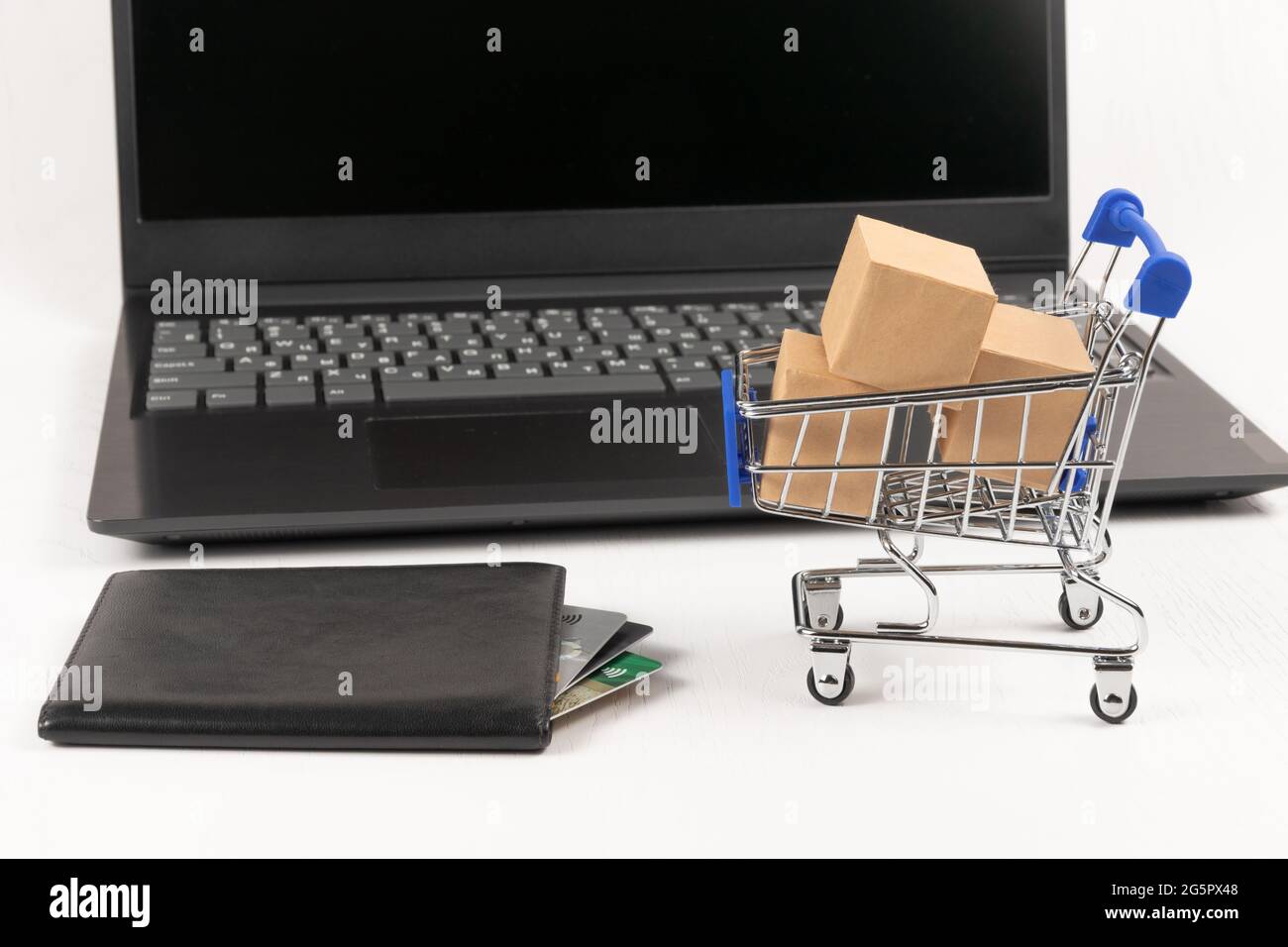 carrello in metallo cromato con scatole e portafoglio in pelle con carte bancarie sullo sfondo di un portatile nero su un tavolo in legno bianco. fuoco selettivo Foto Stock