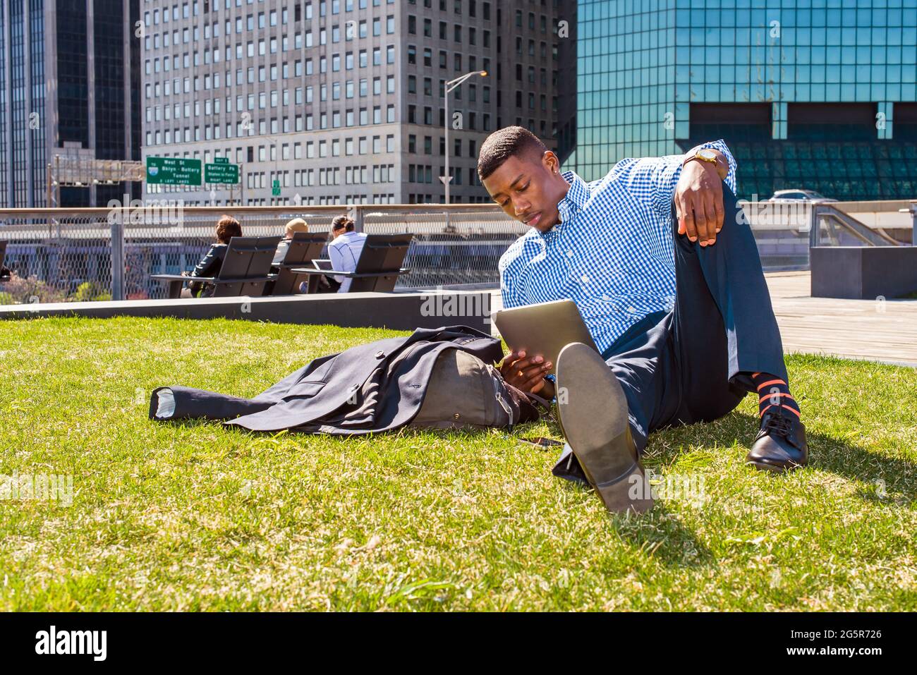 Studiare all'esterno. Un giovane uomo nero è seduto su un prato verde a New York City, leggendo su un computer tablet. Foto Stock