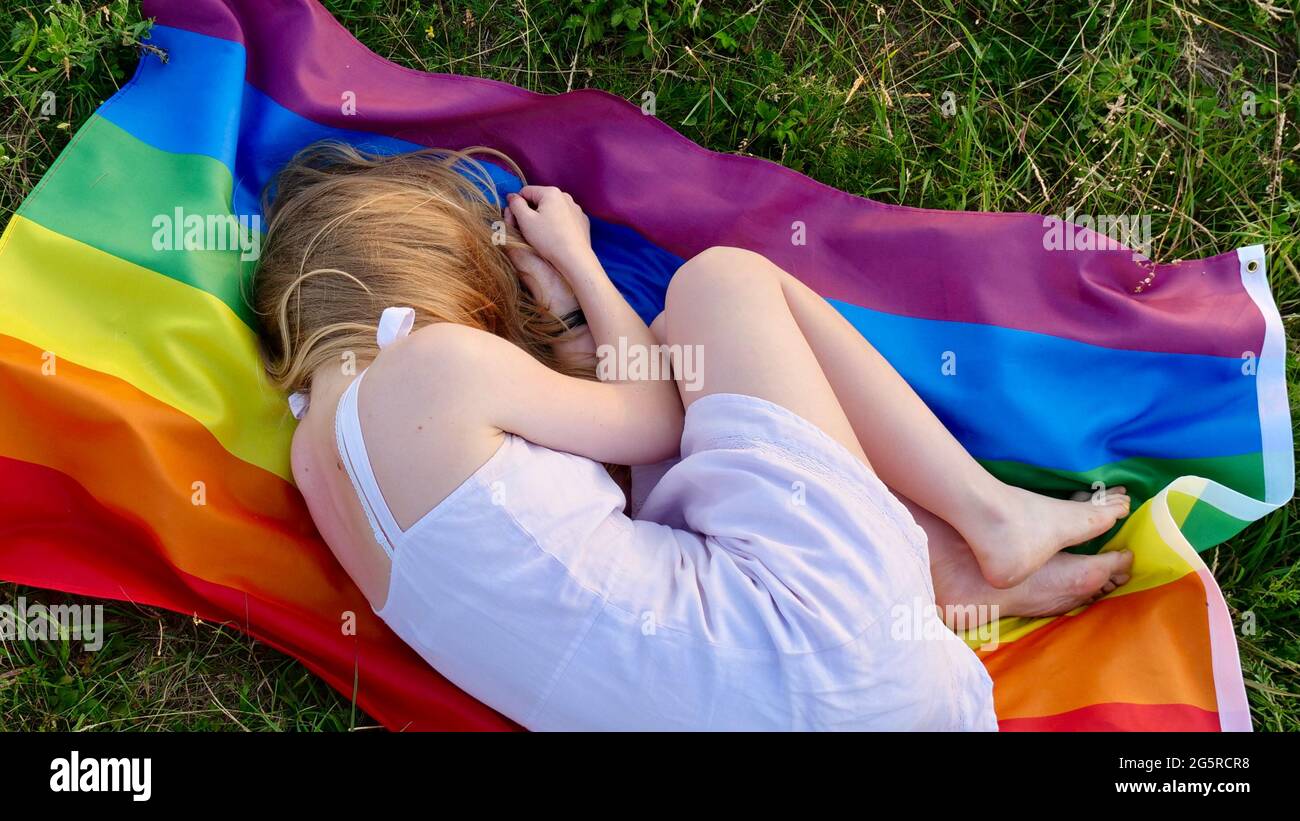 Donna lesbica sdraiata sulla bandiera LGBT sull'erba in abito bianco. Sostegno all'orientamento non tradizionale nel mese della dignità, giorno della bisessualità. La persona perde e nasconde il suo volto dal bullismo. Foto Stock