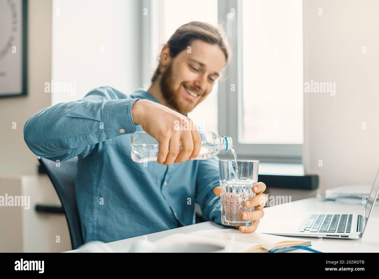 Giovane uomo d'affari felice che versa acqua in vetro sul suo posto di lavoro Foto Stock