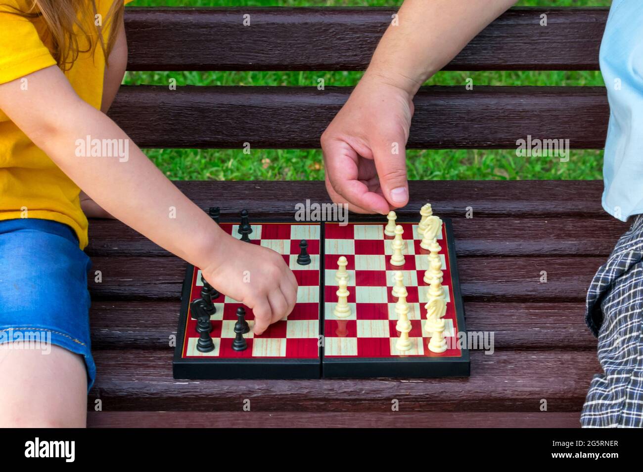 La bambina gioca a scacchi con il papà nel parco su una panchina all'aria aperta. Giorno degli Scacchi. Sviluppo, giochi educativi. Foto Stock