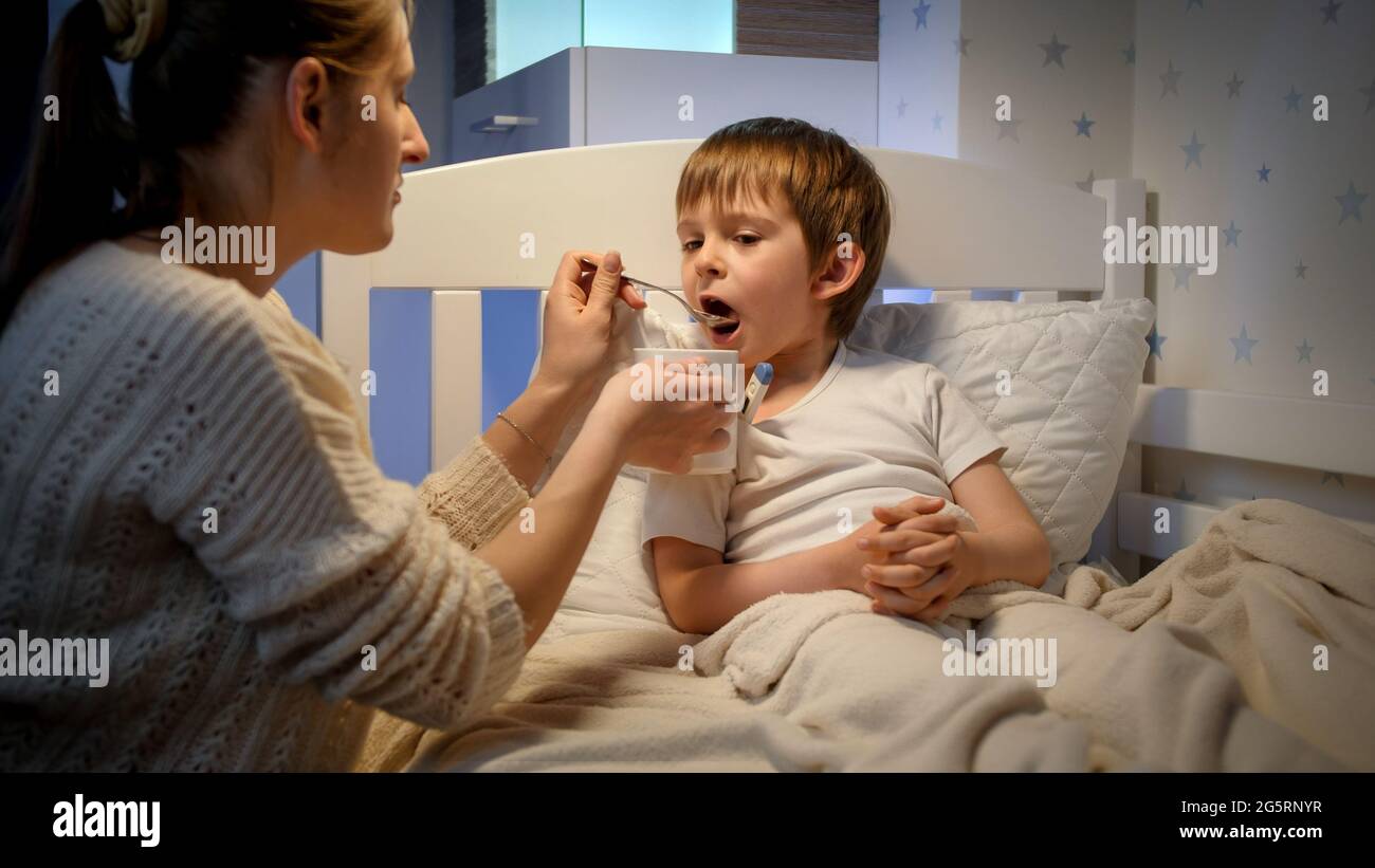 Giovane madre che dà medicine e sciroppo dal cucchiaio al suo figlio malato sdraiato a letto. Concetto di malattia dei bambini, malattia e cura dei genitori Foto Stock