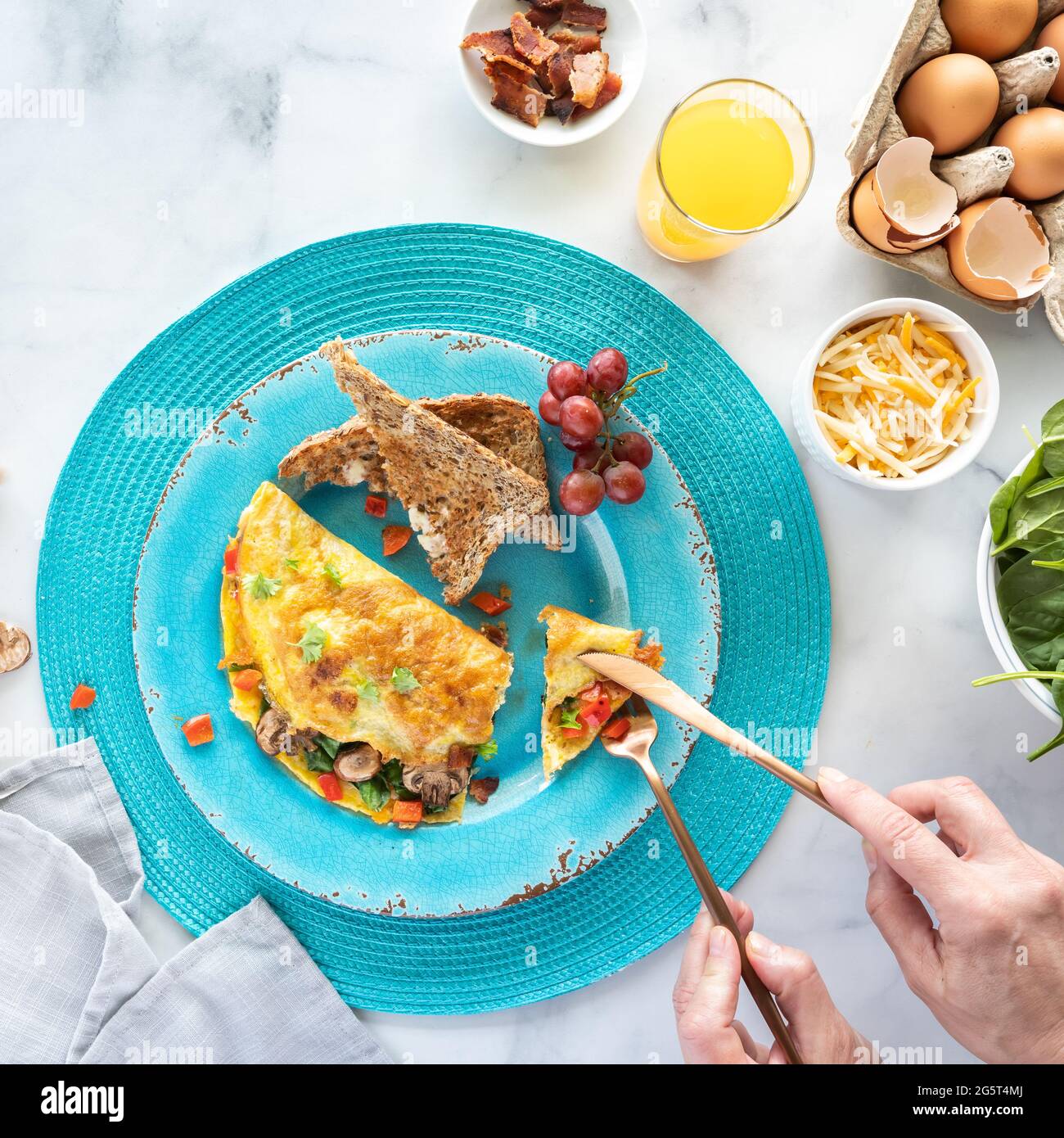 Omelette appena preparate servite con pane tostato e succo d'arancia con le mani usando le posate per tagliare un pezzo. Foto Stock