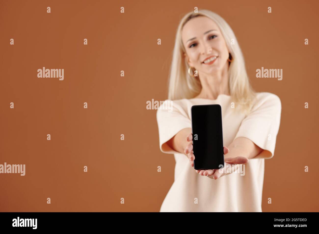 Una giovane donna dai capelli biondi in tshirt che ti dà un nuovo smartphone con schermo vuoto, scatto in studio Foto Stock
