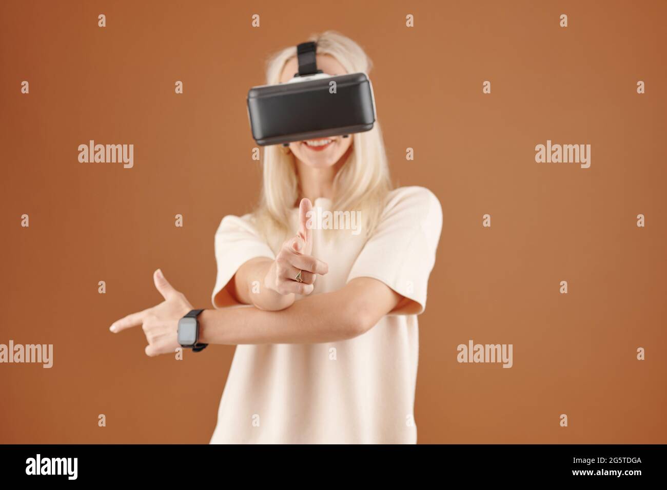 Allegra giovane donna dai capelli biondi in smartwatch che mostra le pistole mentre si gioca a gioco di realtà virtuale su sfondo marrone Foto Stock