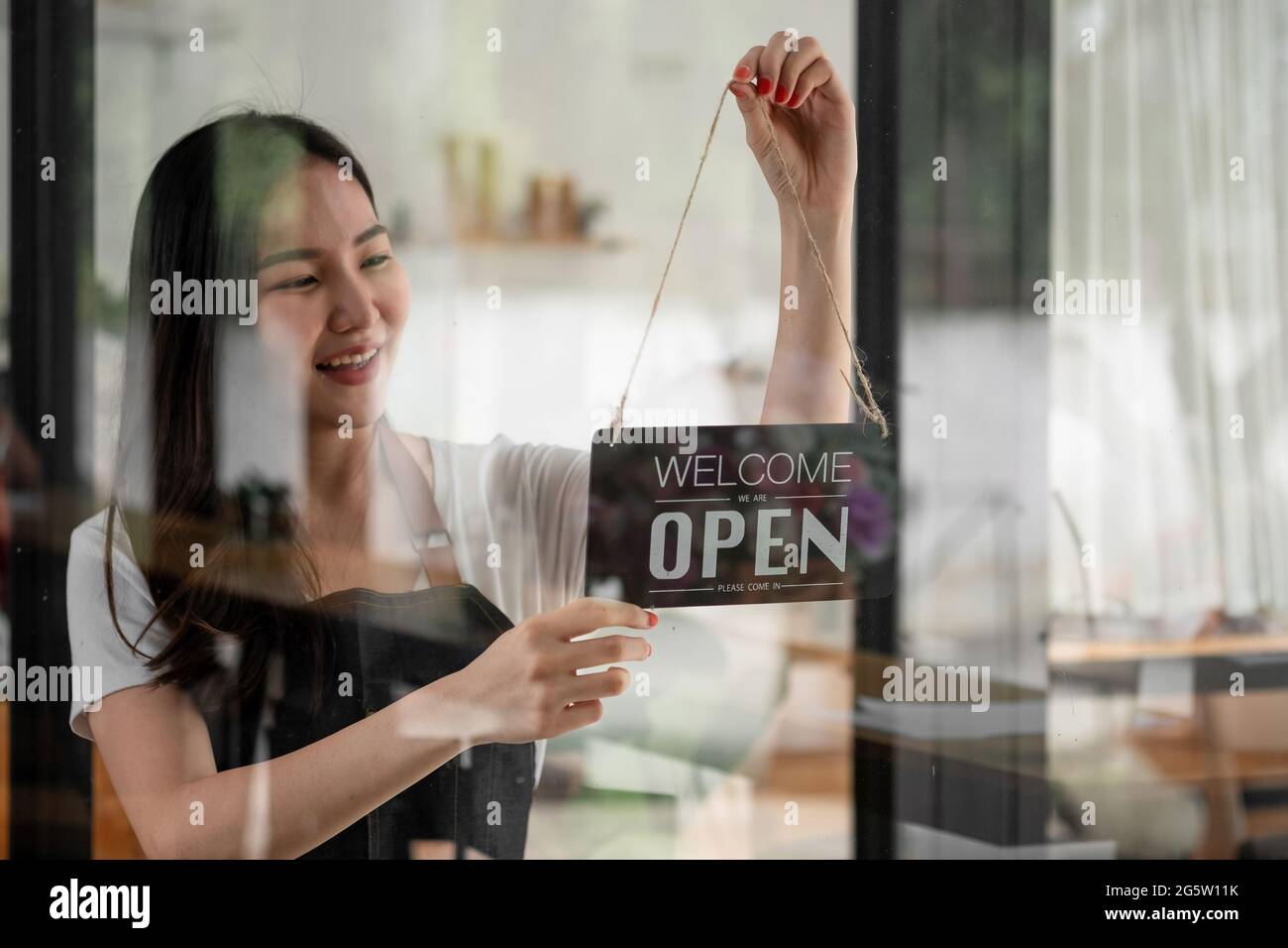Ritratto di una giovane barista sorridente in grembiule che tiene un cartello aperto mentre si trova al suo caffè. Elegante caffetteria asiatica personale femminile girare porta Foto Stock