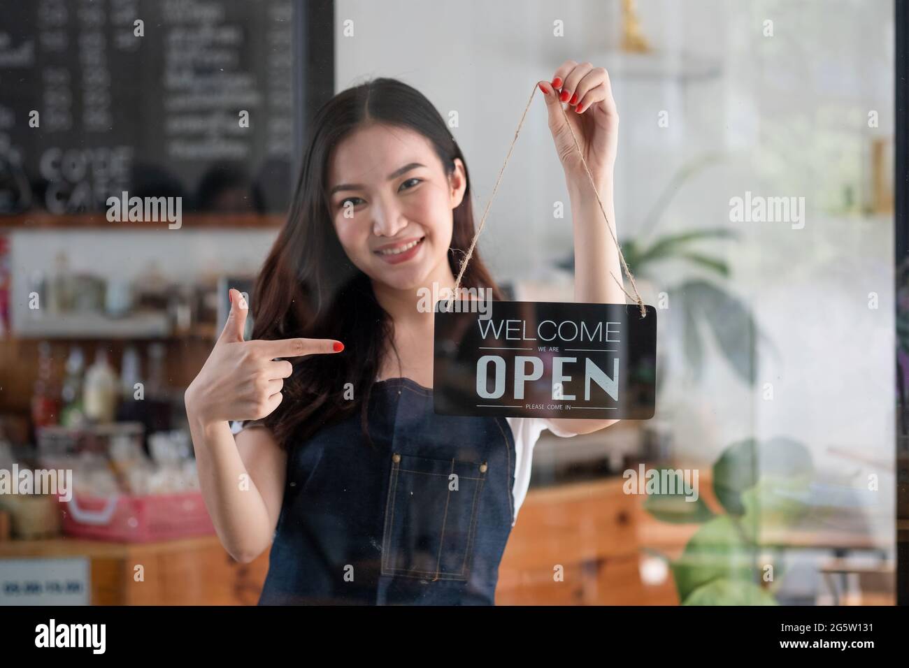 Ritratto di una giovane barista sorridente in grembiule che tiene un cartello aperto mentre si trova al suo caffè. Elegante caffetteria asiatica personale femminile girare porta Foto Stock