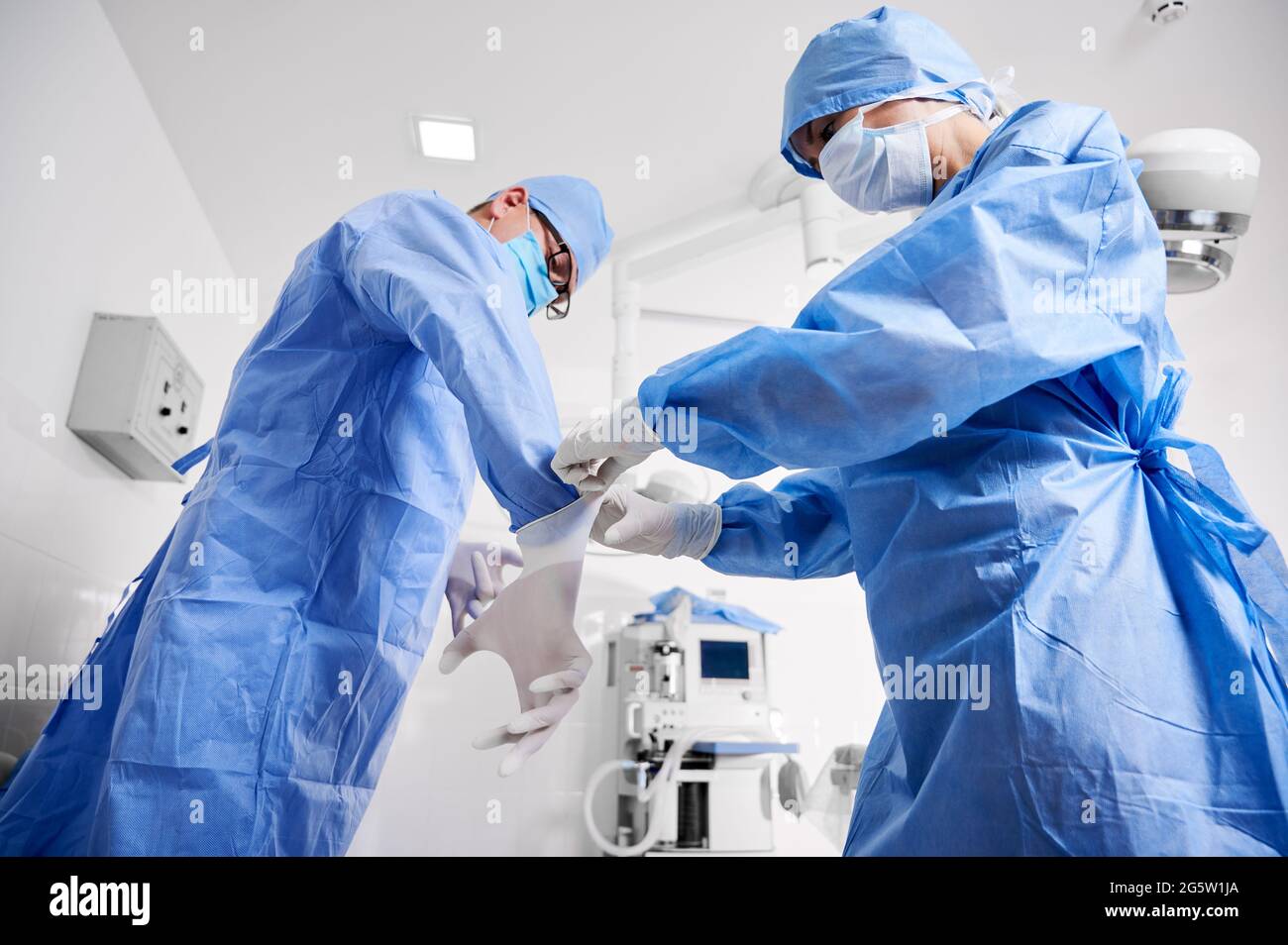 Vista laterale dell'assistente in tuta chirurgica blu che aiuta il chirurgo a indossare guanti sterili. Due medici in maschere mediche protettive che si preparano per la chirurgia plastica in clinica. Concetto di medicina e chirurgia. Foto Stock