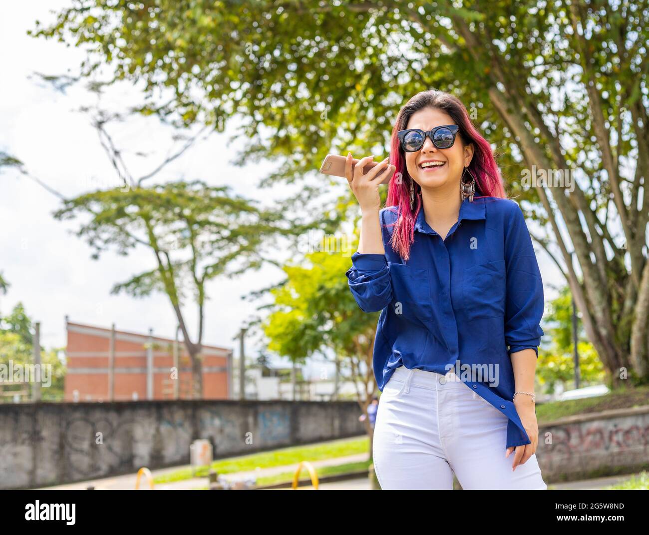 Giovane latina sorridente felice ascolto messaggio audio utilizzando smartphone al parco Foto Stock