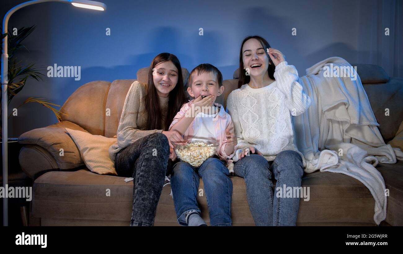 Ragazzo sorridente con due ragazze adolescenti che guardano lo spettacolo televisivo di tarda notte e mangiano il popcorn dalla grande ciotola Foto Stock