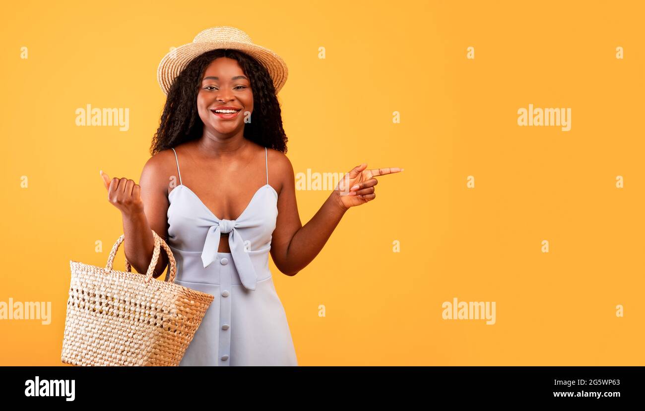 Affascinante donna nera in abito e cappello che tiene sacchetto di paglia, che punta da parte a spazio libero su sfondo arancione, banner Foto Stock