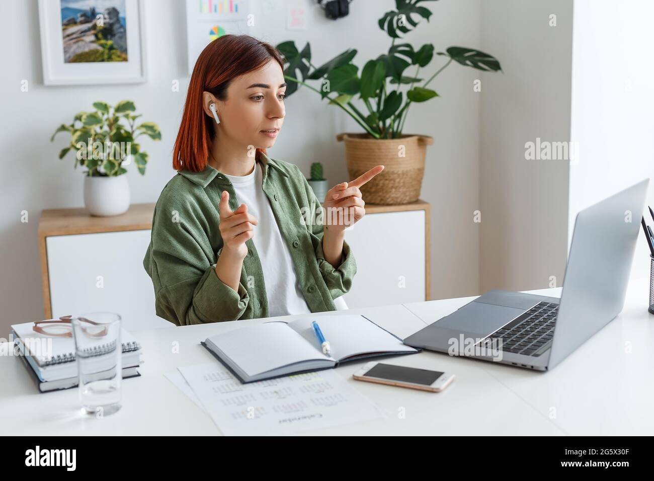 Donna che lavora in remoto a casa con un computer portatile. Giovane donna d'affari che parla con gli auricolari wireless. Concetto di business online o di comunicazione, freelance Foto Stock