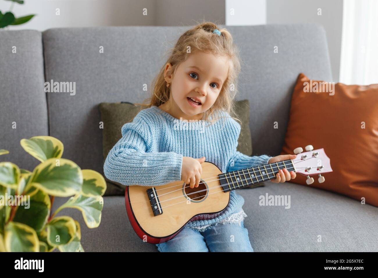 Ritratto di carina bambina che gioca ukulele e lo canta a casa. 3 anni bambino imparare chitarra. Concetto di educazione della prima infanzia, hobbistica musicale Foto Stock