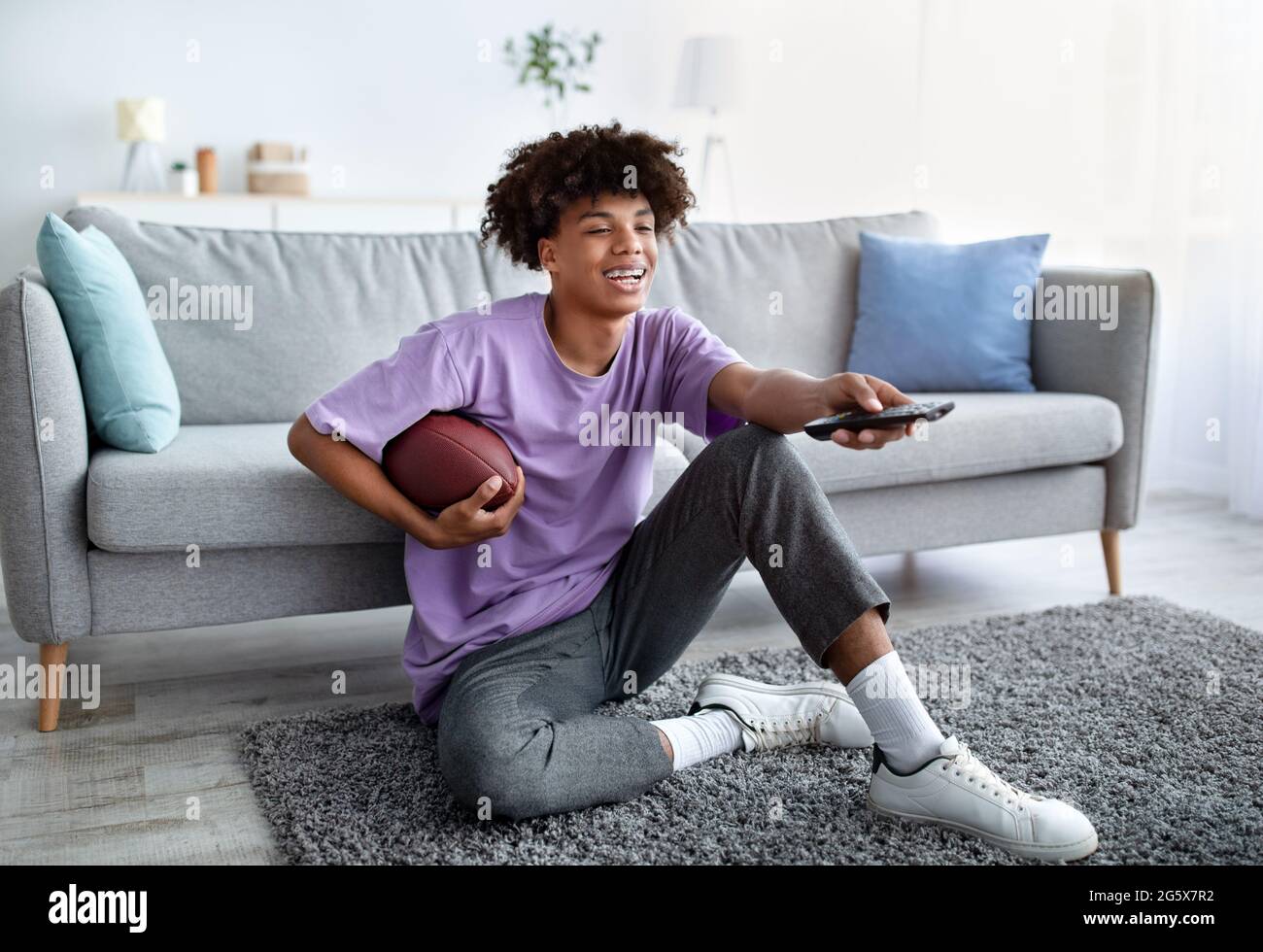 Un ragazzo teen nero che guarda il calcio americano in TV, tenendo il telecomando e la palla a casa Foto Stock