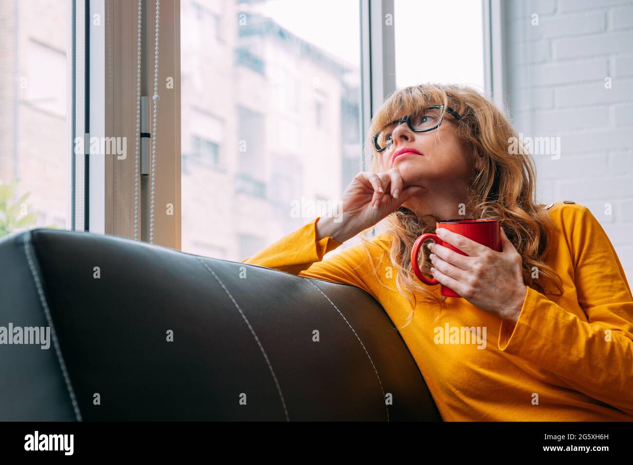 ritratto di una donna adulta pensiva o preoccupata a casa guardando fuori la finestra con una tazza di caffè Foto Stock