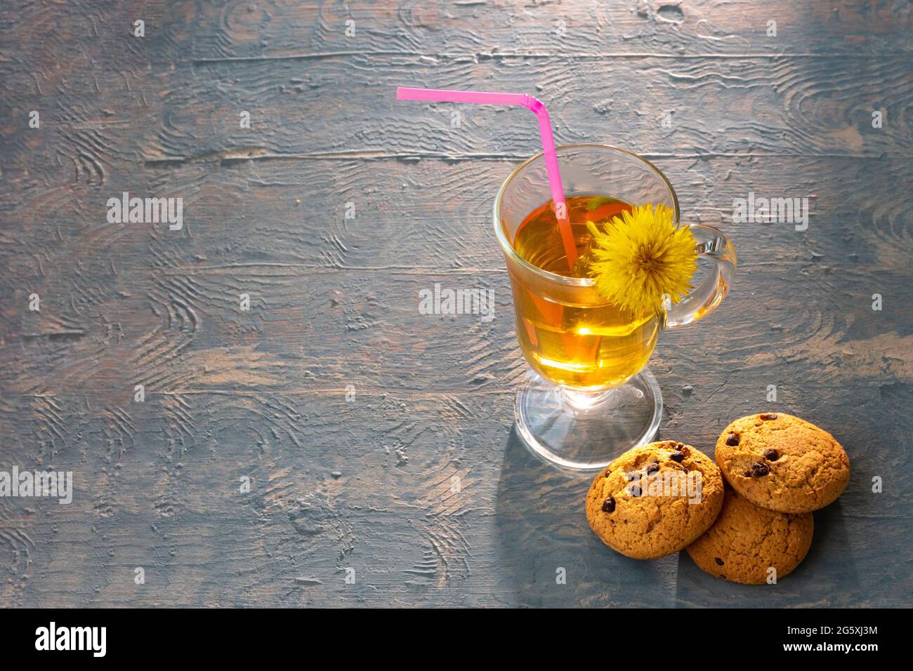 Tazza di tè con tè alle erbe, dente di leone giallo e paglia rosa e biscotti con uvetta su sfondo di legno blu illuminato da raggi solari, spazio copia. Hea Foto Stock