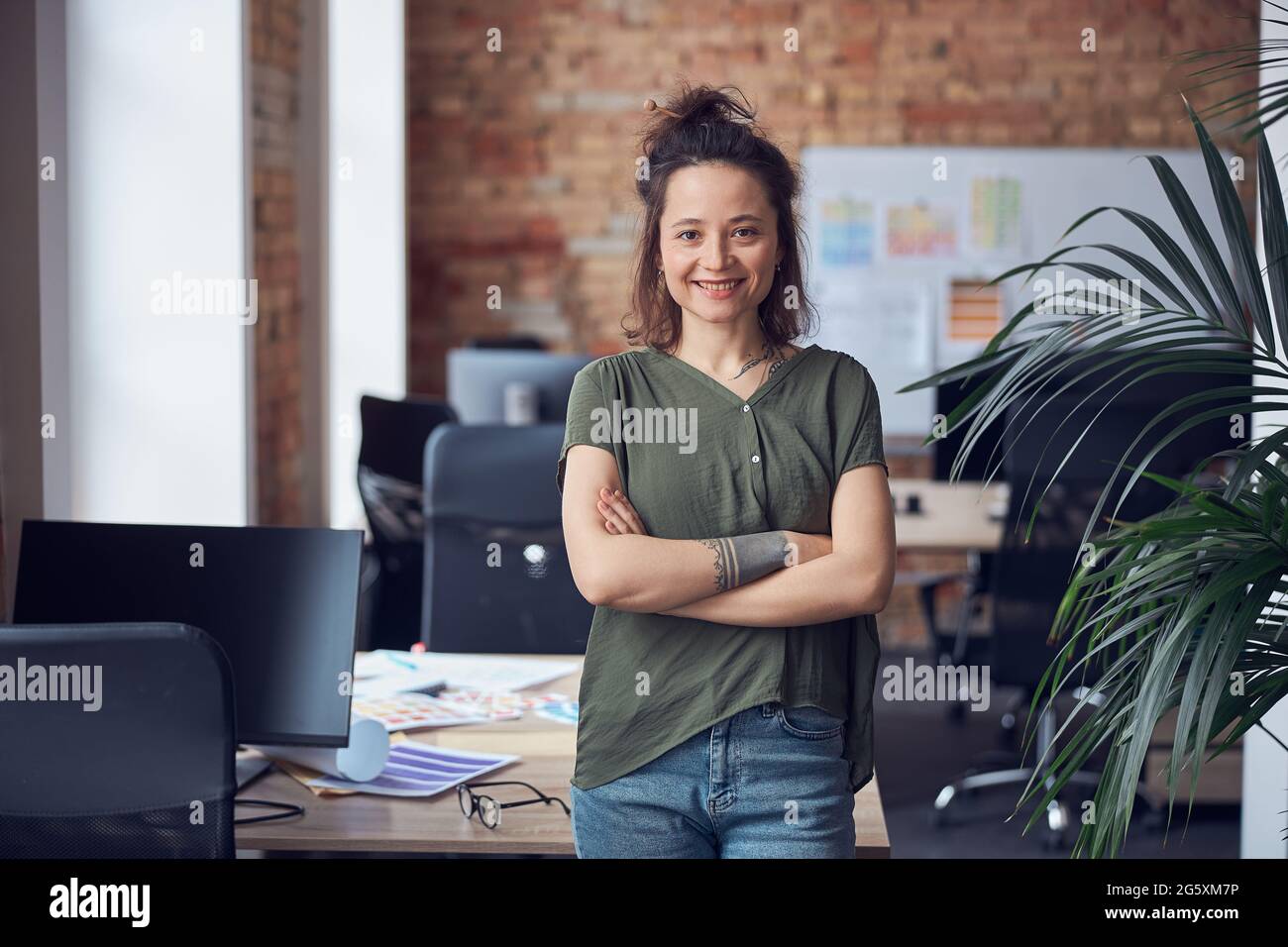 Ritratto di donna felice, designer di interni o architetto sorridente alla macchina fotografica mentre in piedi con le braccia incrociate in un moderno ufficio verde di giorno Foto Stock
