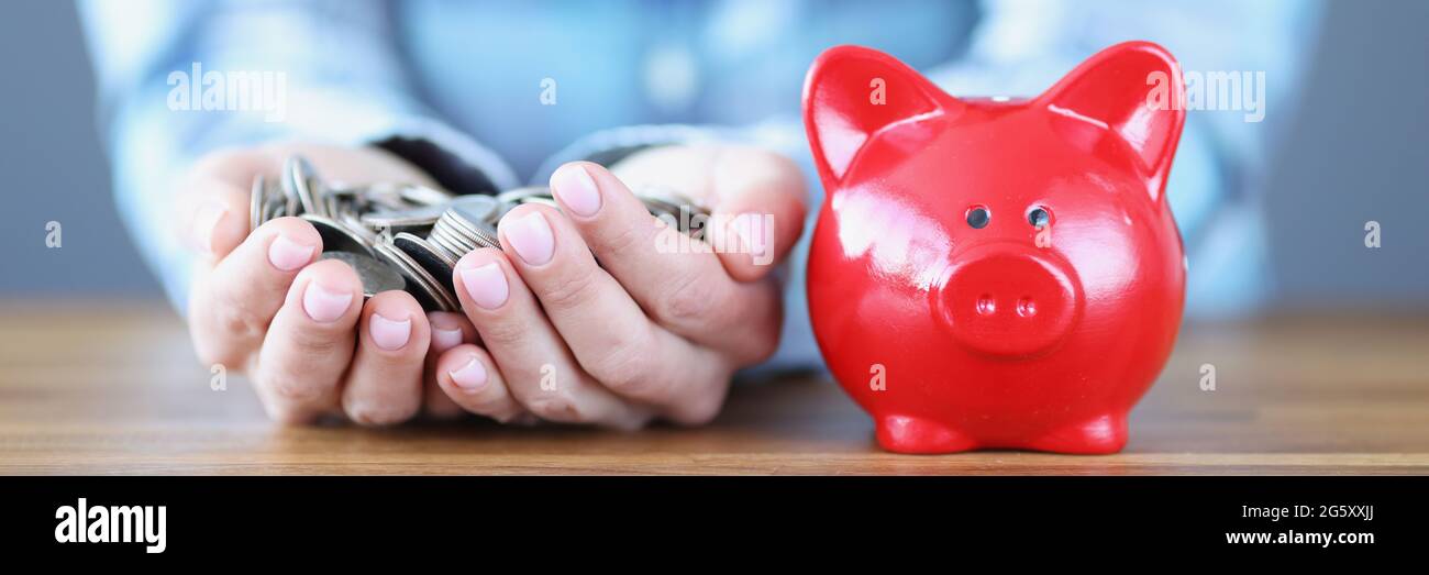 Le mani femminili stanno tenendo manciate di monete vicino al rosso porcellino banca Foto Stock