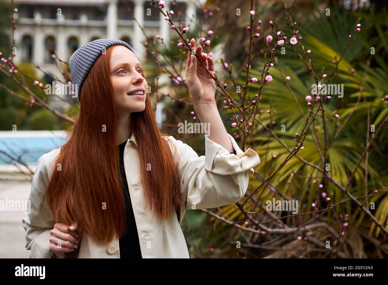 Adorabile donna dai capelli rossi in cappello che si pone in fiore albero a primavera. Bella giovane donna felice godendo l'odore nel giardino fiorente della sorgente, toccando fl Foto Stock